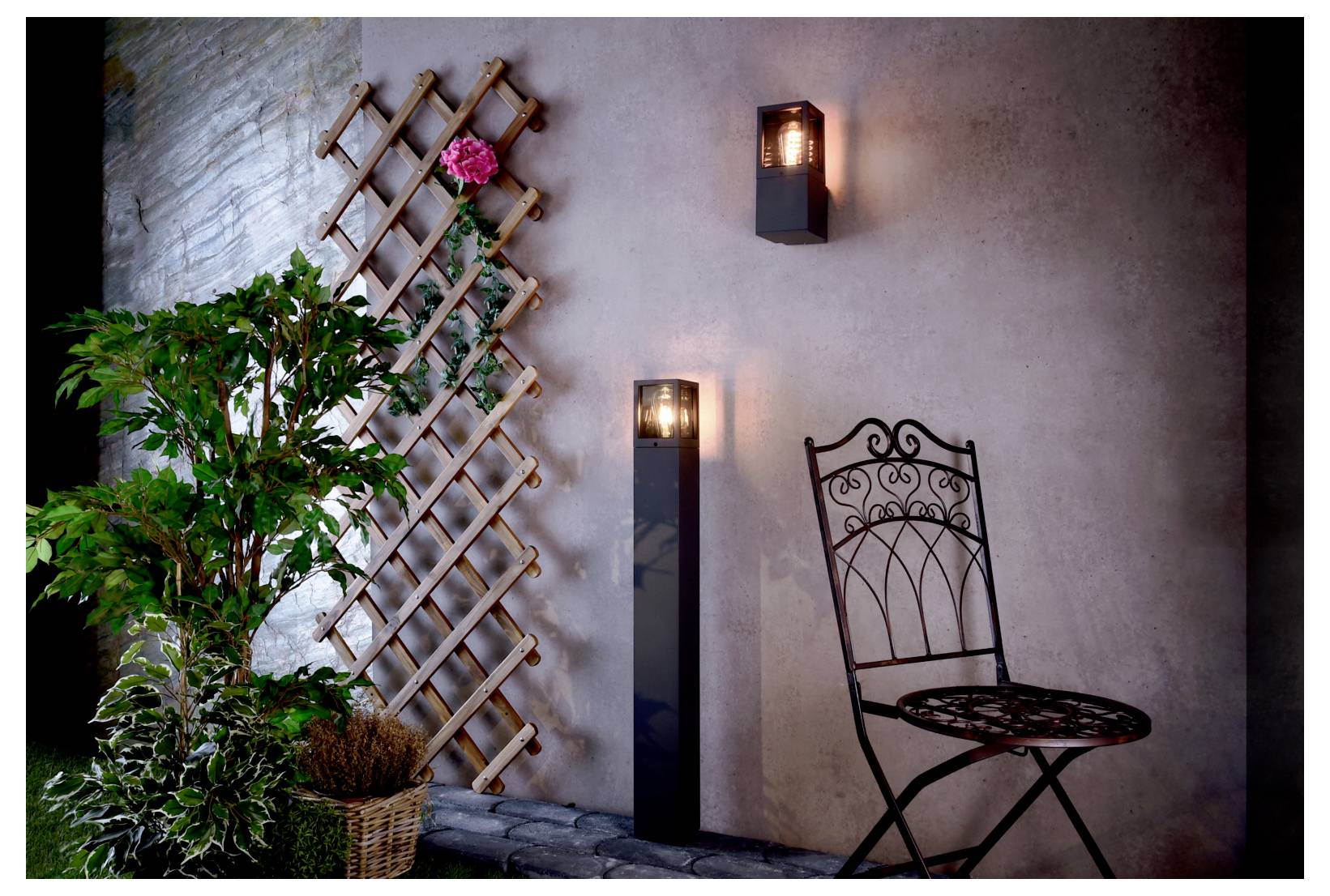 Terrassenecke mit Stuhl aus Schmiedeeisen, Wandleuchten, Gitterspalier mit rosa Blüten und Topfpflanzen vor einer Stuckwand.