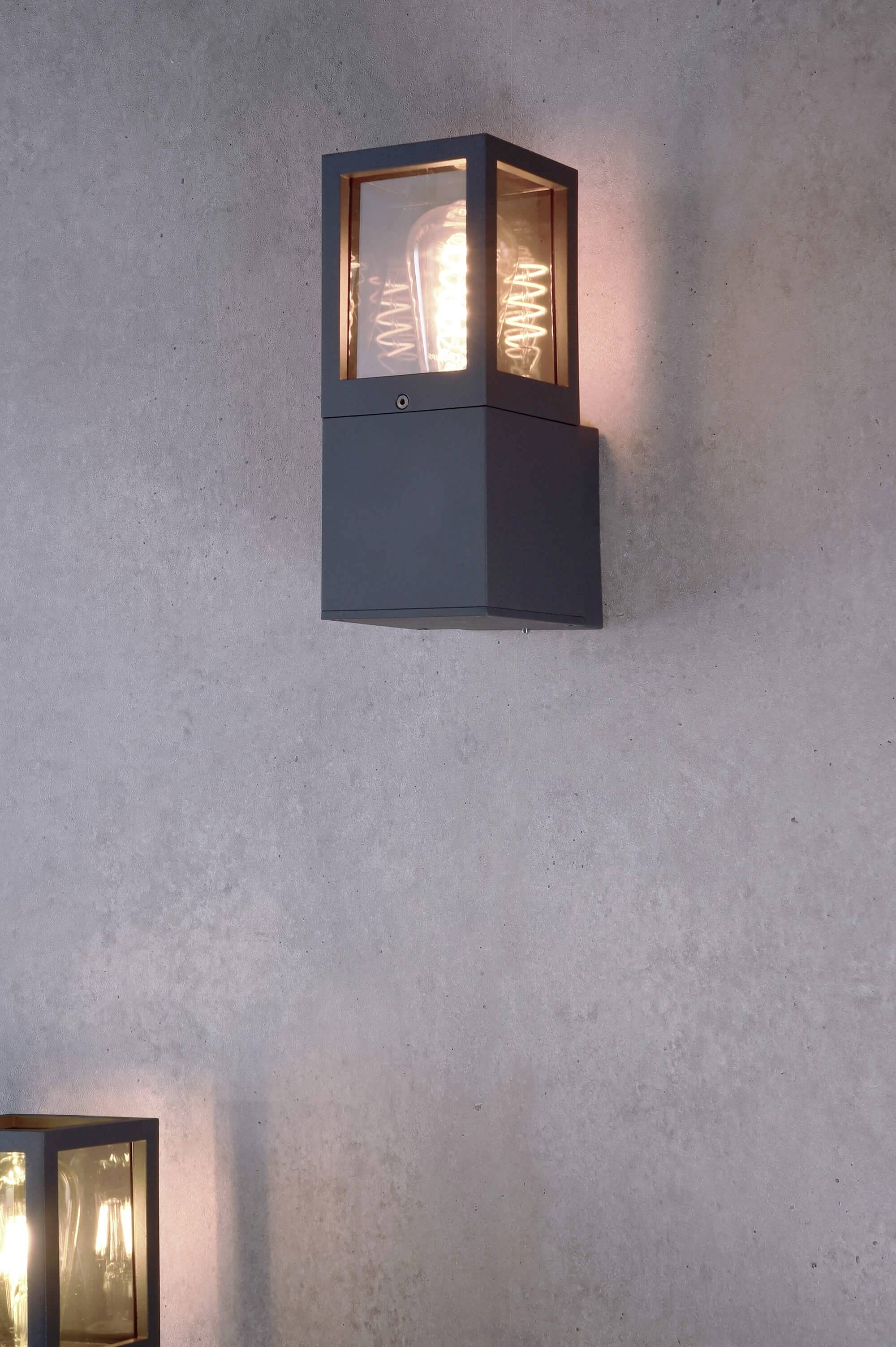 Eine moderne Wandleuchte mit eckigem, transparentem Gehäuse und spiraliger Glühbirne, montiert an einer grauen Betonwand.