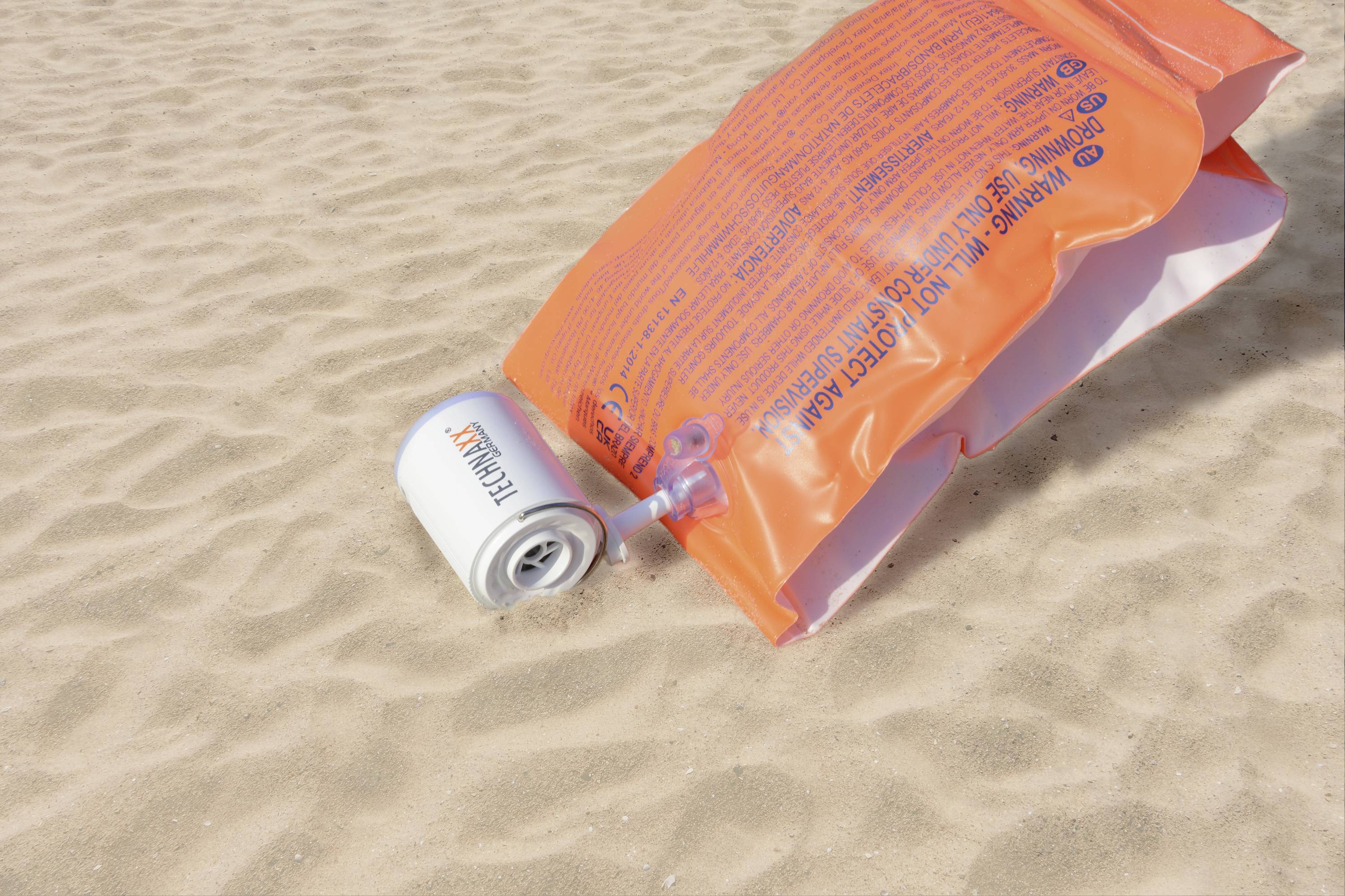 Eine orangefarbene Schwimmweste und eine Thermoskanne liegen auf sandigem Boden am Strand.