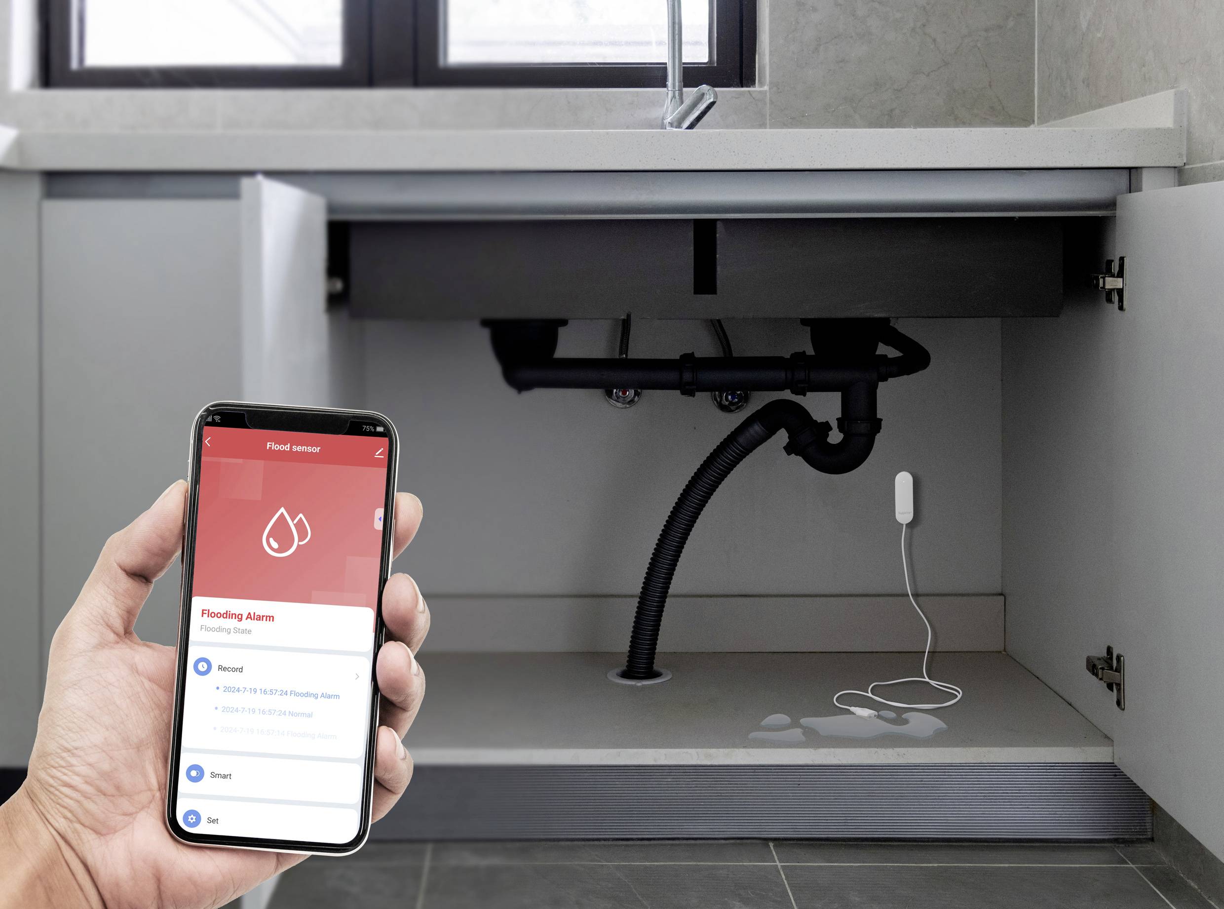 Eine Hand hält ein Smartphone mit einer App, die einen Überschwemmungsalarm zeigt. Im Hintergrund ein Wassersensor unter einer Spüle.