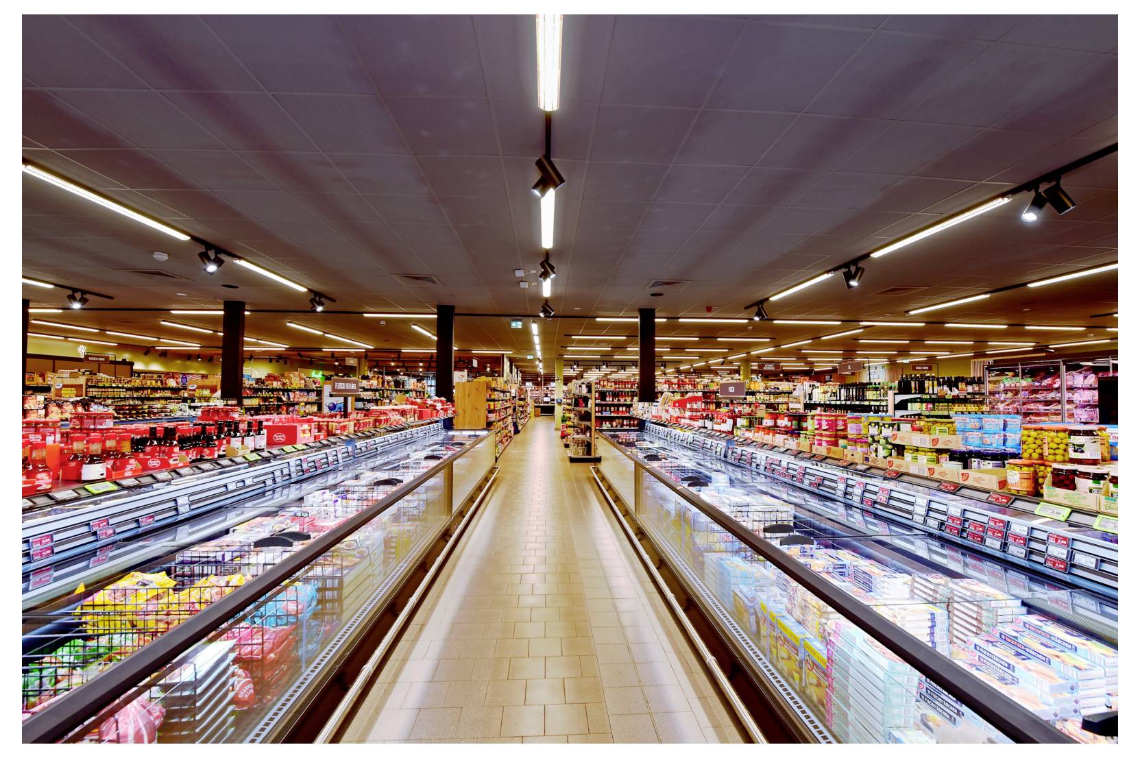 Breiter Supermarktgang mit Kühlvitrinen auf beiden Seiten und Regalen voller Lebensmittel im Hintergrund.