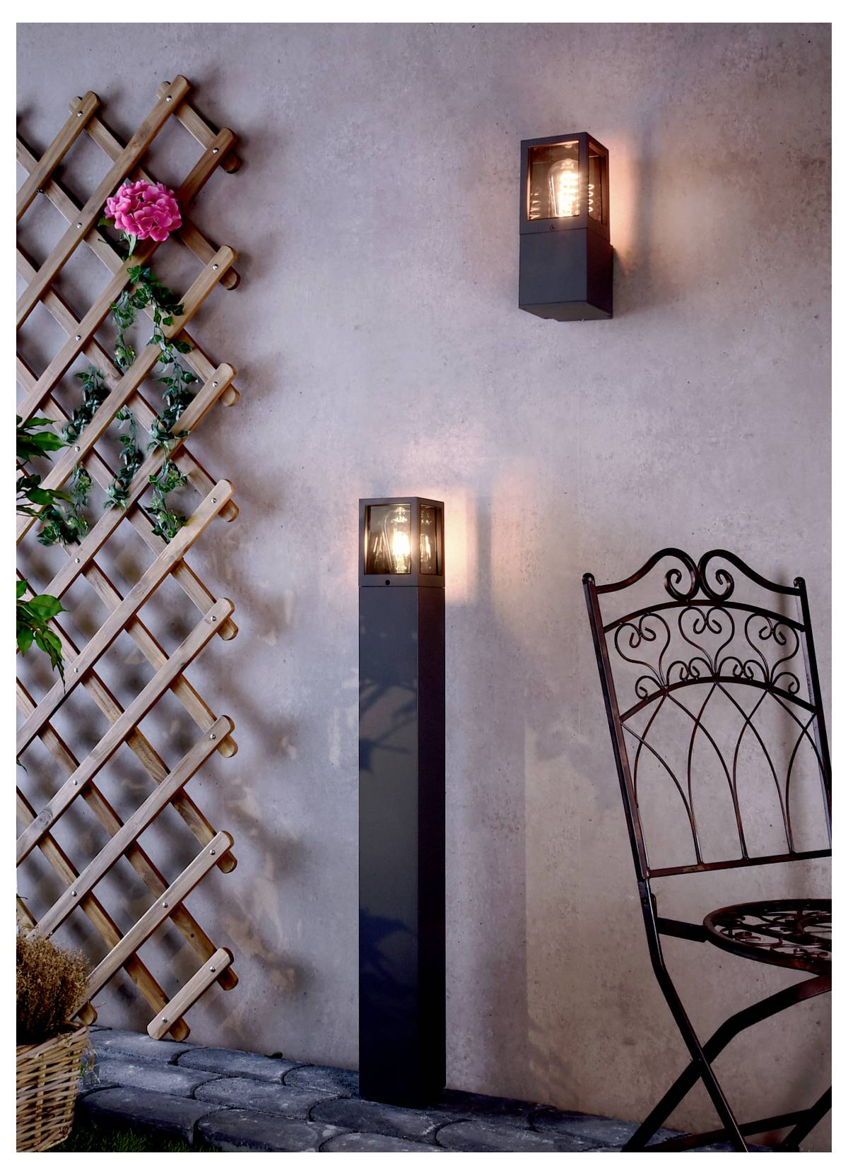 Außenwand mit zwei beleuchteten Wandleuchten, einem Holzspalier mit rosa Blüten und einem Teil eines verzierten Metallstuhls auf einer Steinterrasse.