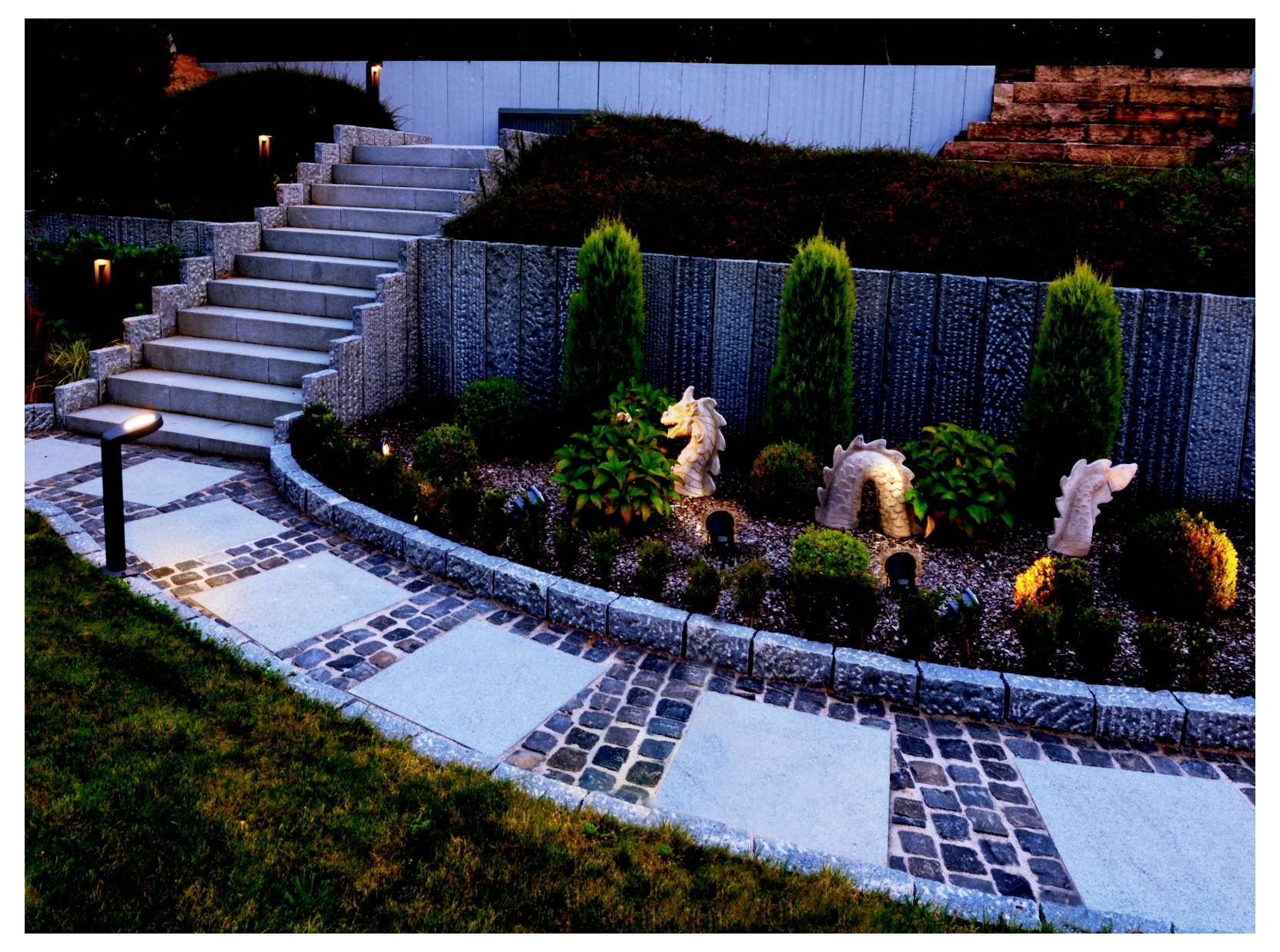 Geschwungener Gartenweg neben Steinstufen, mit kleinen beleuchteten Statuen und Sträuchern in einem gestalteten Hinterhof in der Dämmerung.