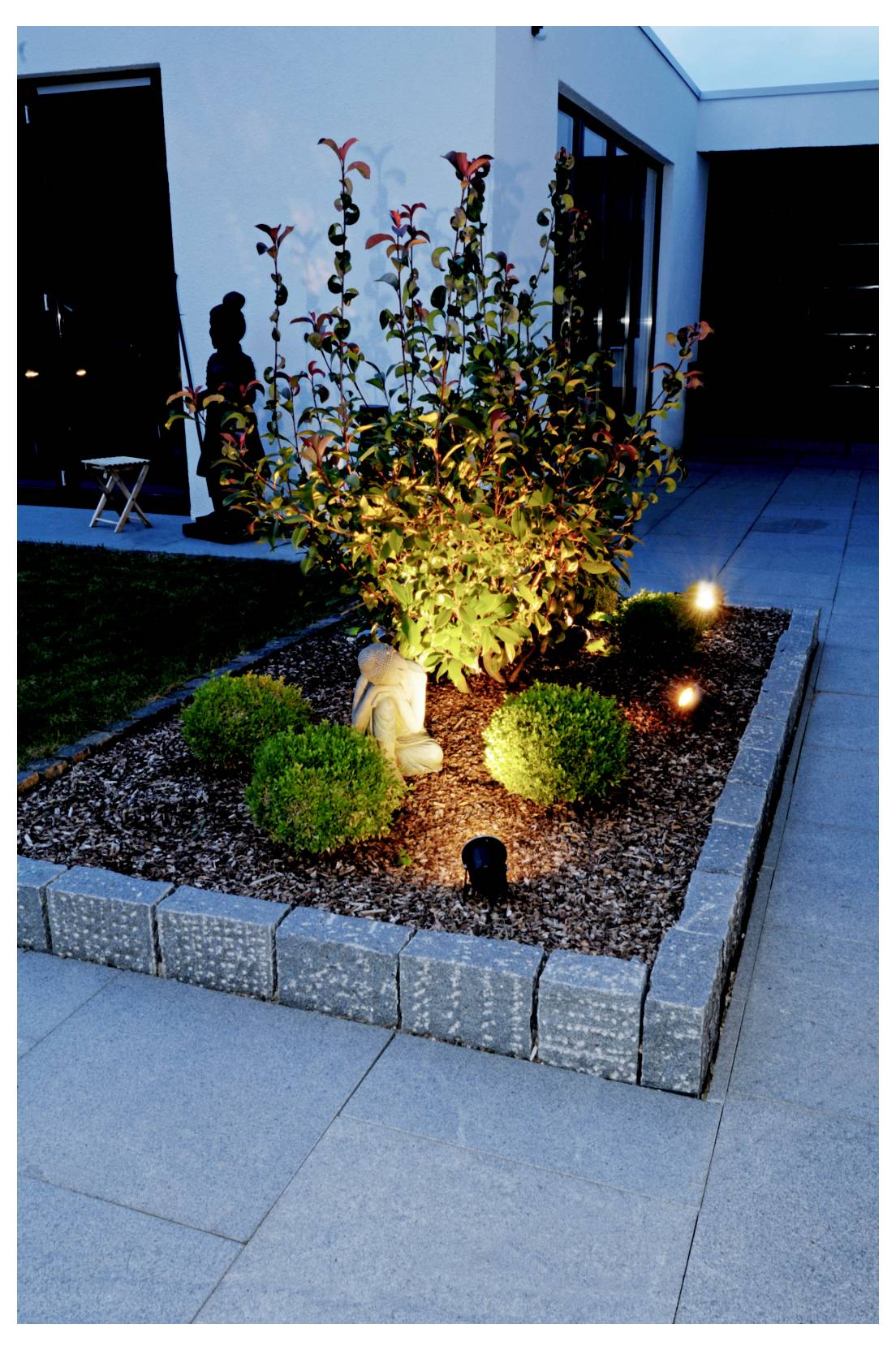 Beleuchtetes, gestaltetes Gartenbeet mit einem kleinen Zierbaum, niedrigen Sträuchern und Bodenleuchten neben einer modernen Hausterrasse in der Dämmerung.