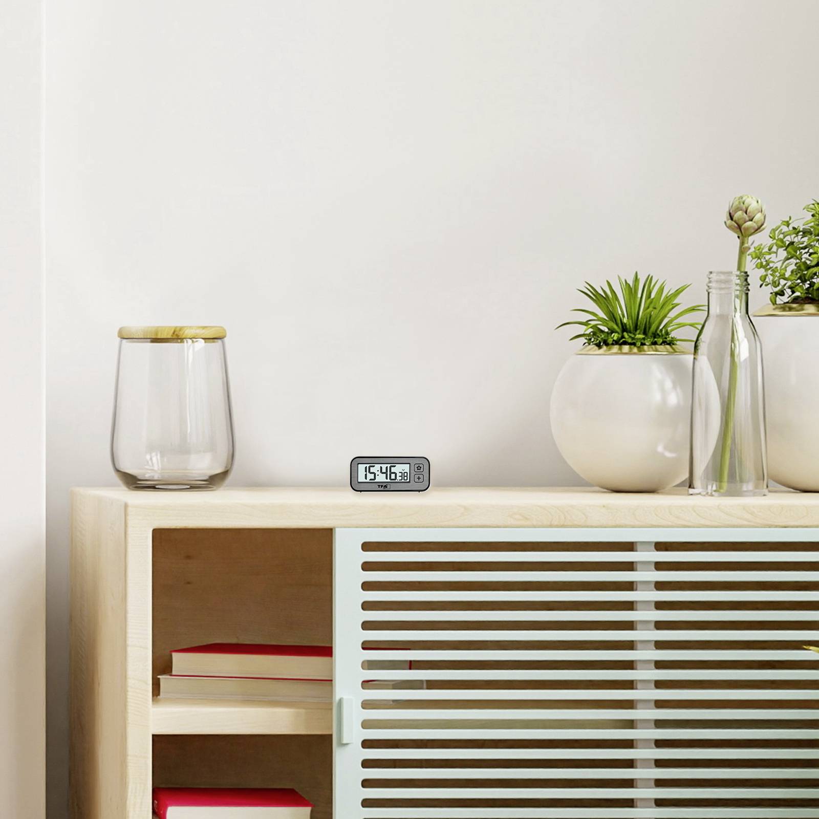 Ein Holztisch mit einer digitalen Uhr, grüner Pflanze in einem Glasbehälter und zwei Bücher im Regal darunter.