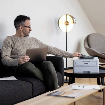Ein Mann sitzt auf einem Sofa und bedient einen Laptop. Neben ihm steht ein Drucker auf einem Tisch. Im Hintergrund ist eine Stehlampe.
