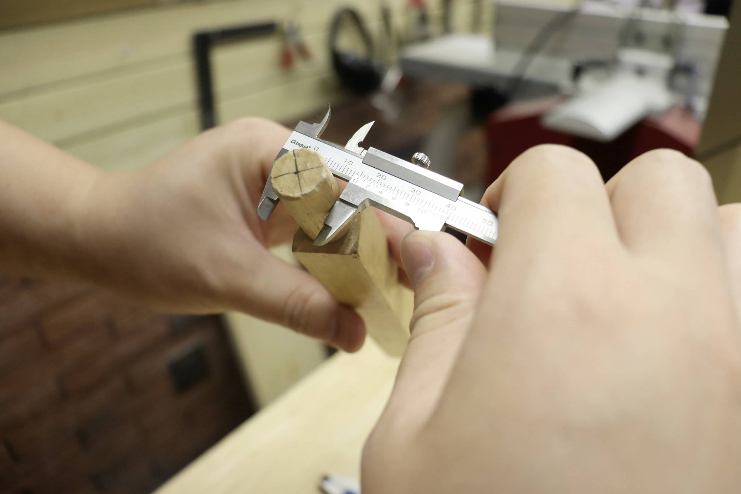 Zwei Hände messen mit einem Messschieber den Durchmesser eines Holzstücks in einer Werkstatt.