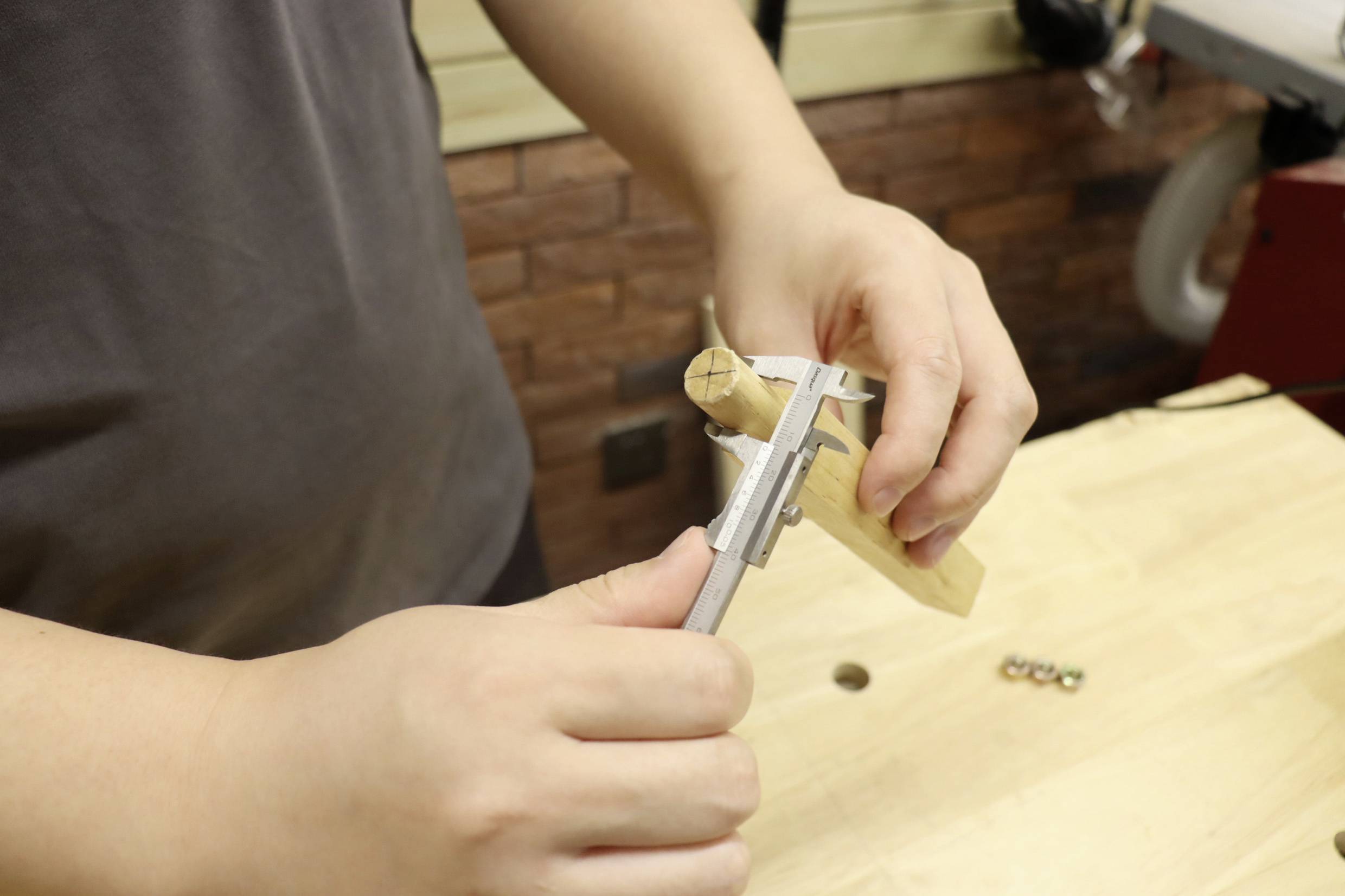 Eine Person misst ein rundes Holzstück mit einem Messschieber auf einer Werkbank. Drei Schrauben liegen daneben.