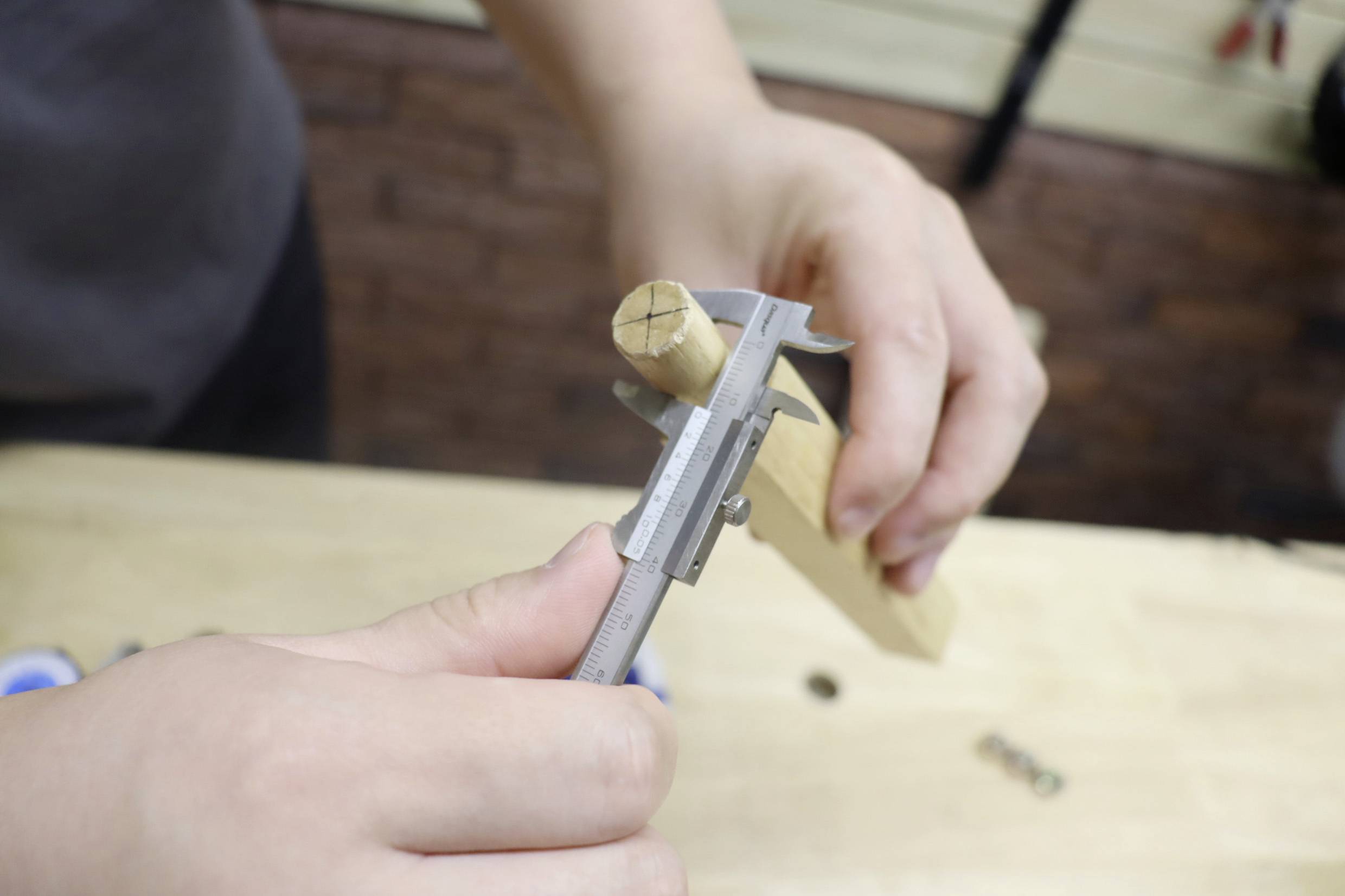 Eine Person misst den Durchmesser eines Holzstabs mit einem Messschieber auf einem Holztisch.
