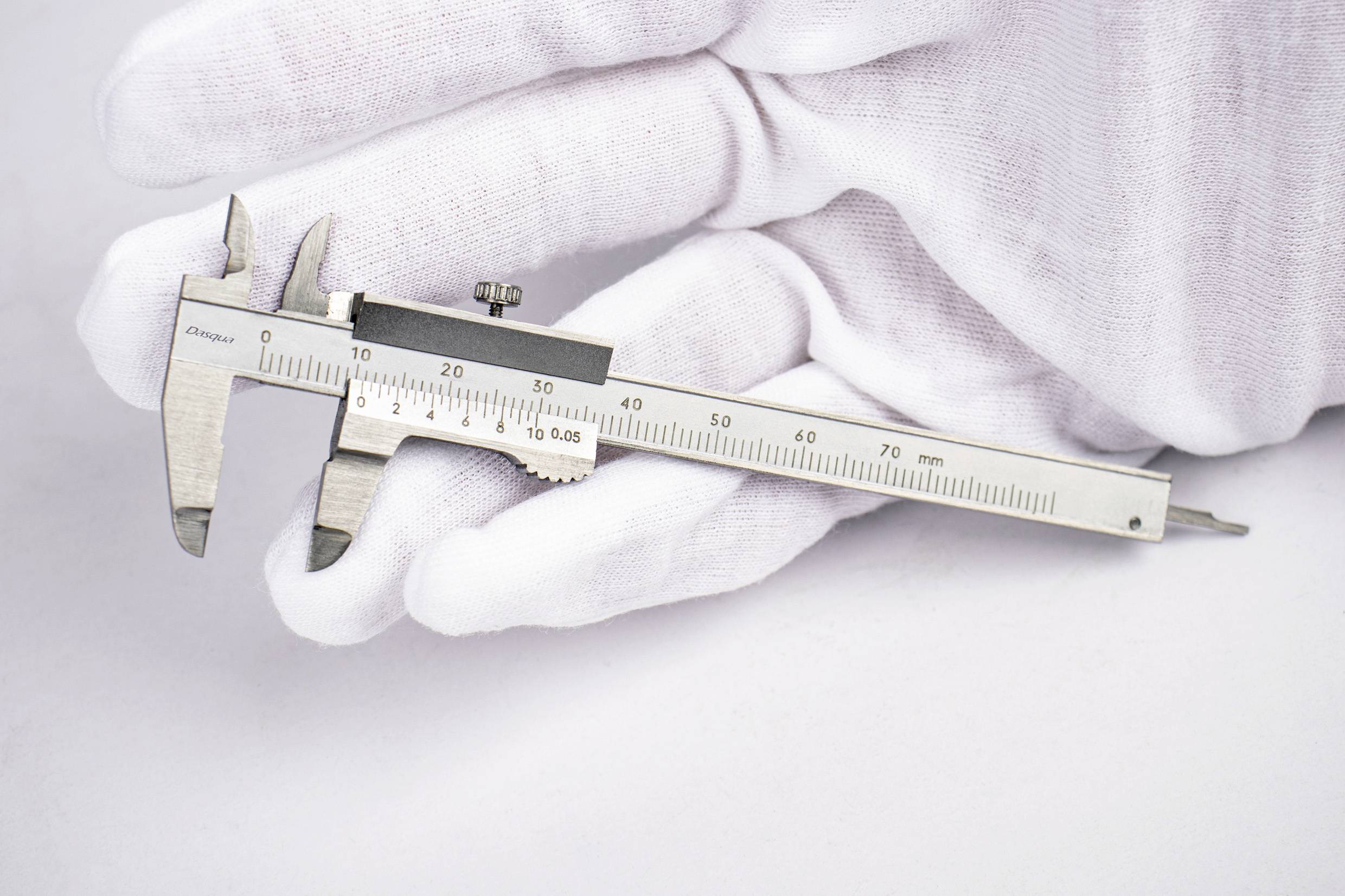 Eine Hand in einem weißen Handschuh hält einen kleinen Messschieber vor einem neutralen Hintergrund.