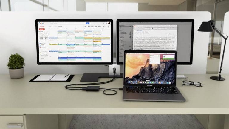 Ein Arbeitsplatz mit einem Laptop und zwei Monitoren, die Kalender- und Dokumentanwendungen anzeigen. Bürobedarf wie Notizbuch und Brille.