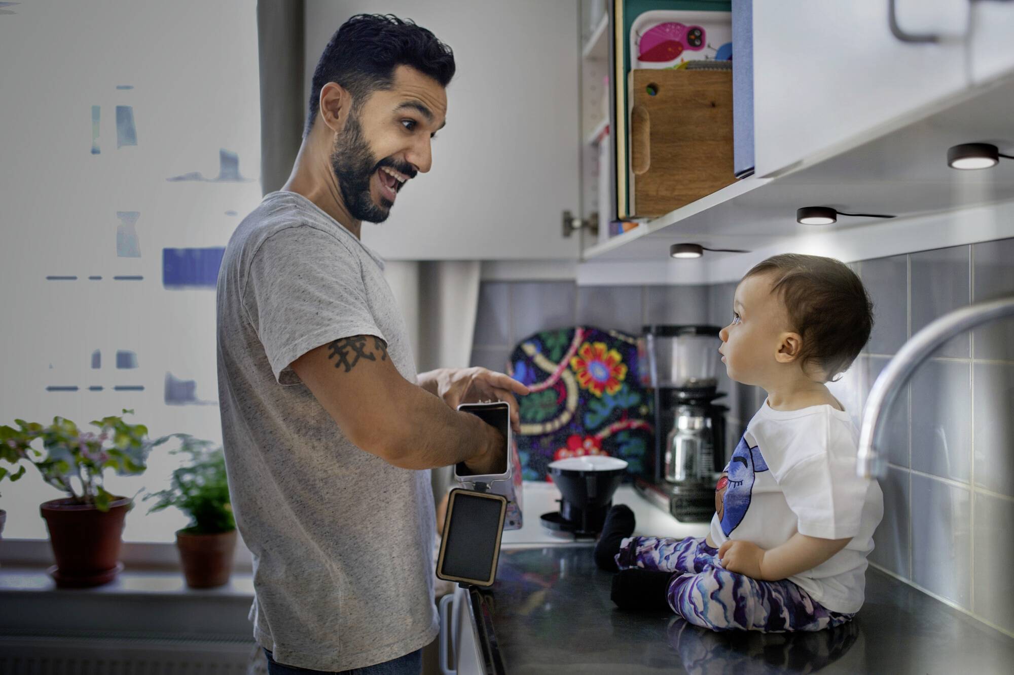 Ein Mann lächelt und spricht mit einem Kleinkind, das auf der Küchentheke sitzt. Sie sind in einer hellen, modernen Küche.