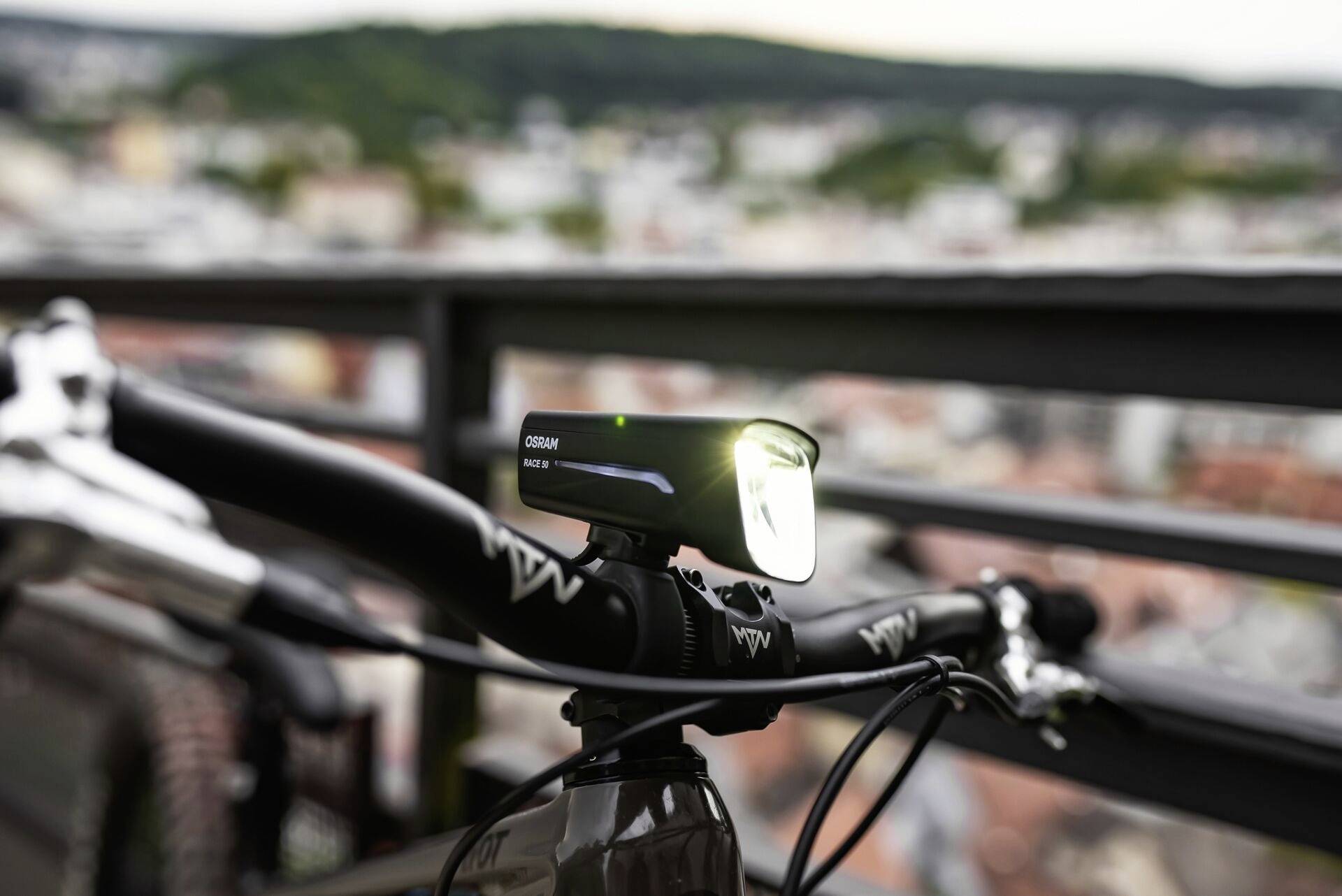 Eine Fahrradleuchte ist an einem Fahrradlenker befestigt. Im Hintergrund ist eine unscharfe Stadtlandschaft sichtbar.