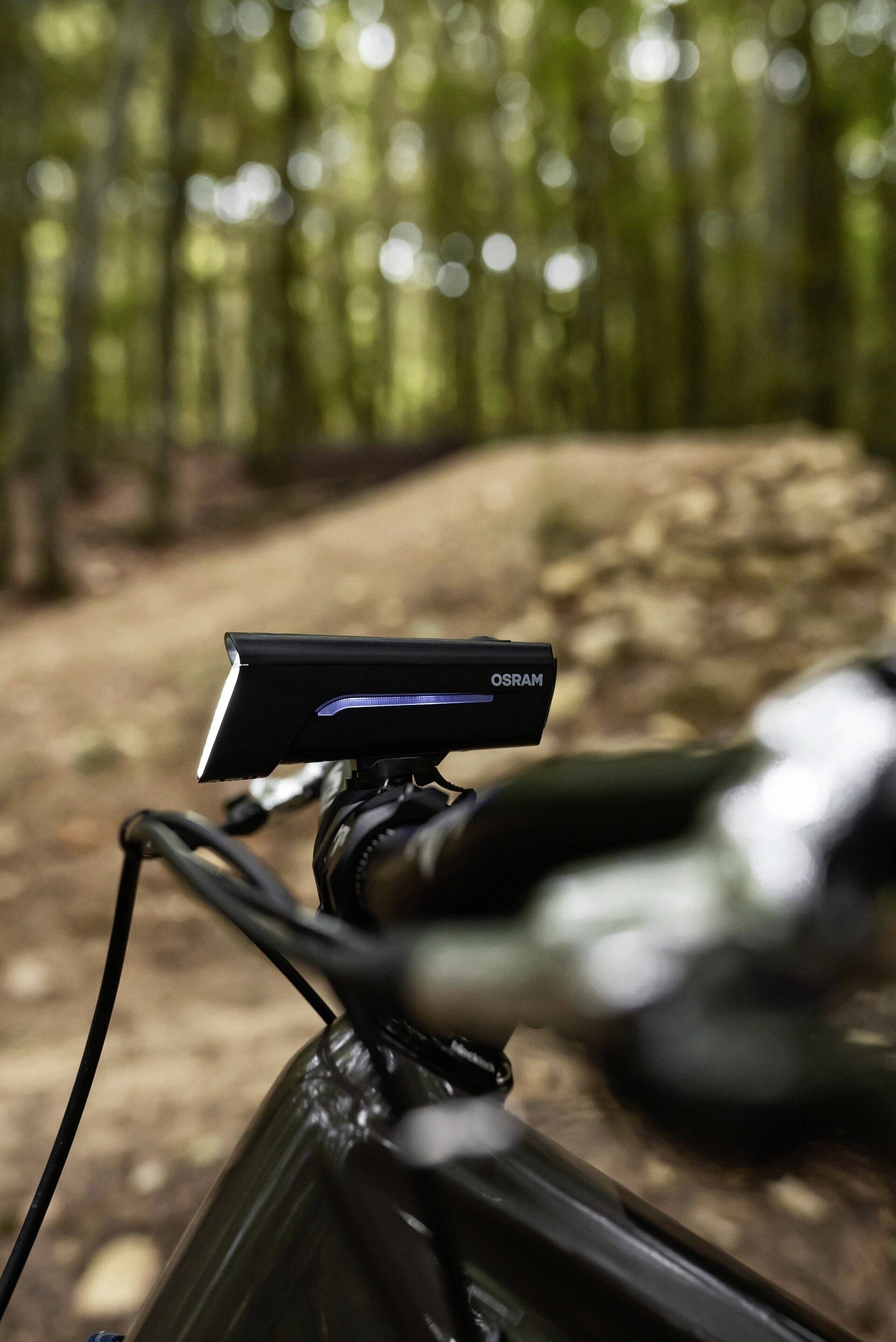 Fahrradlenker mit einem elektronischen Gerät auf einem Waldweg, unscharfer Hintergrund mit Bäumen, vermittelt einen Outdoor-Abenteuer-Eindruck.