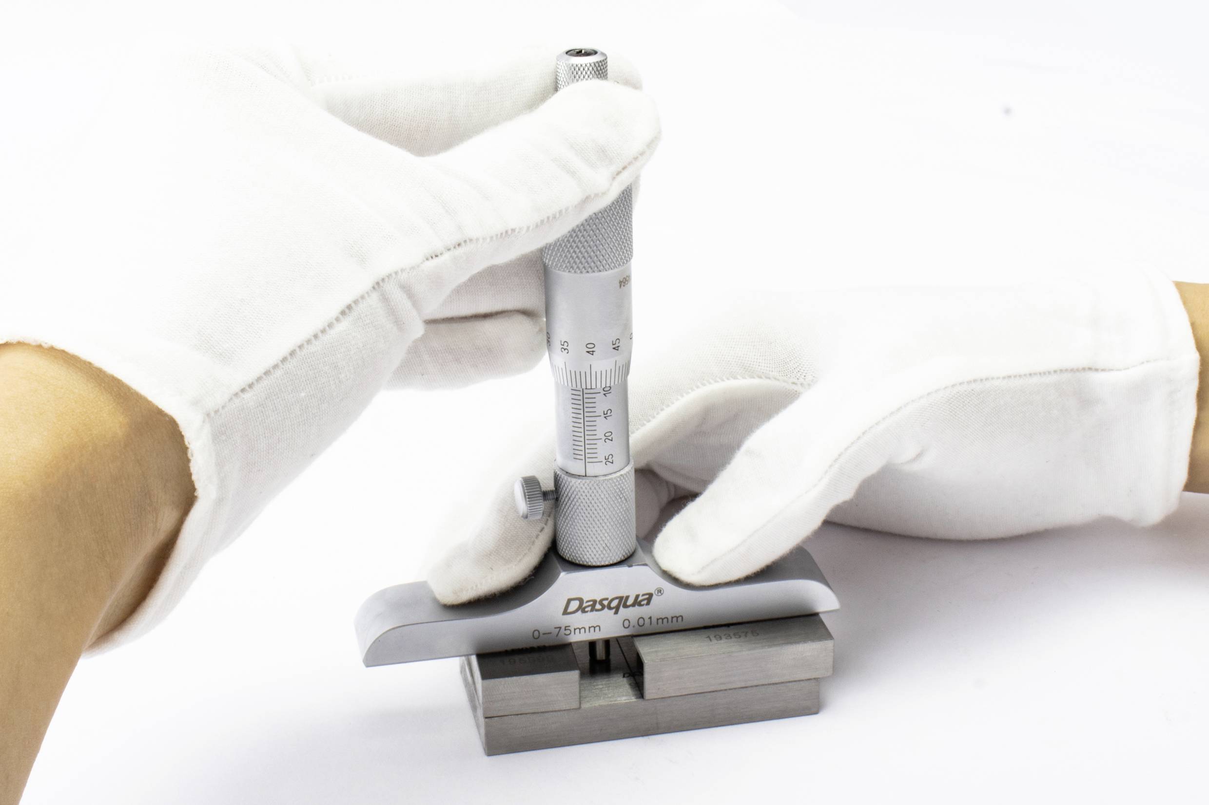Zwei Hände in weißen Handschuhen halten ein Mikrometer, das die Dicke eines Metallblocks misst.