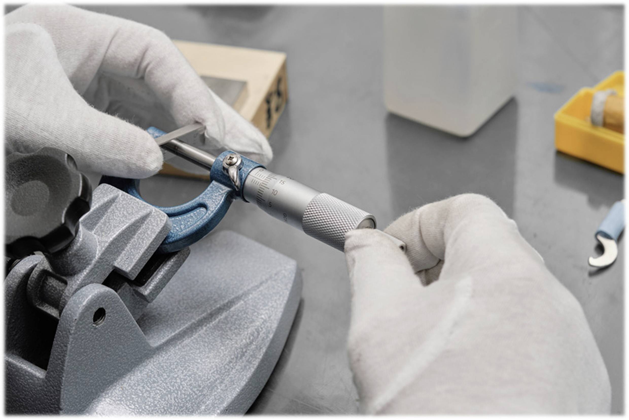 Eine Person misst mit einem Mikrometer und trägt weiße Handschuhe. Weitere Werkzeuge und Geräte sind auf dem Tisch sichtbar.
