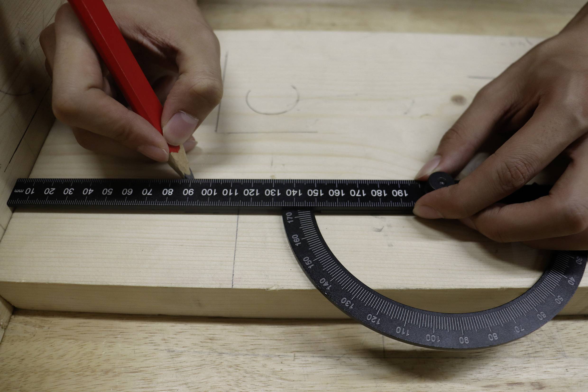 Eine Person zeichnet mit einem Bleistift eine Linie auf ein Holzbrett, wobei sie ein Geodreieck zur genauen Messung verwendet.
