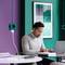 Eine Person arbeitet konzentriert an einem Tisch mit Laptop und Notizbuch in einem modern eingerichteten Büro mit grüner Wand.