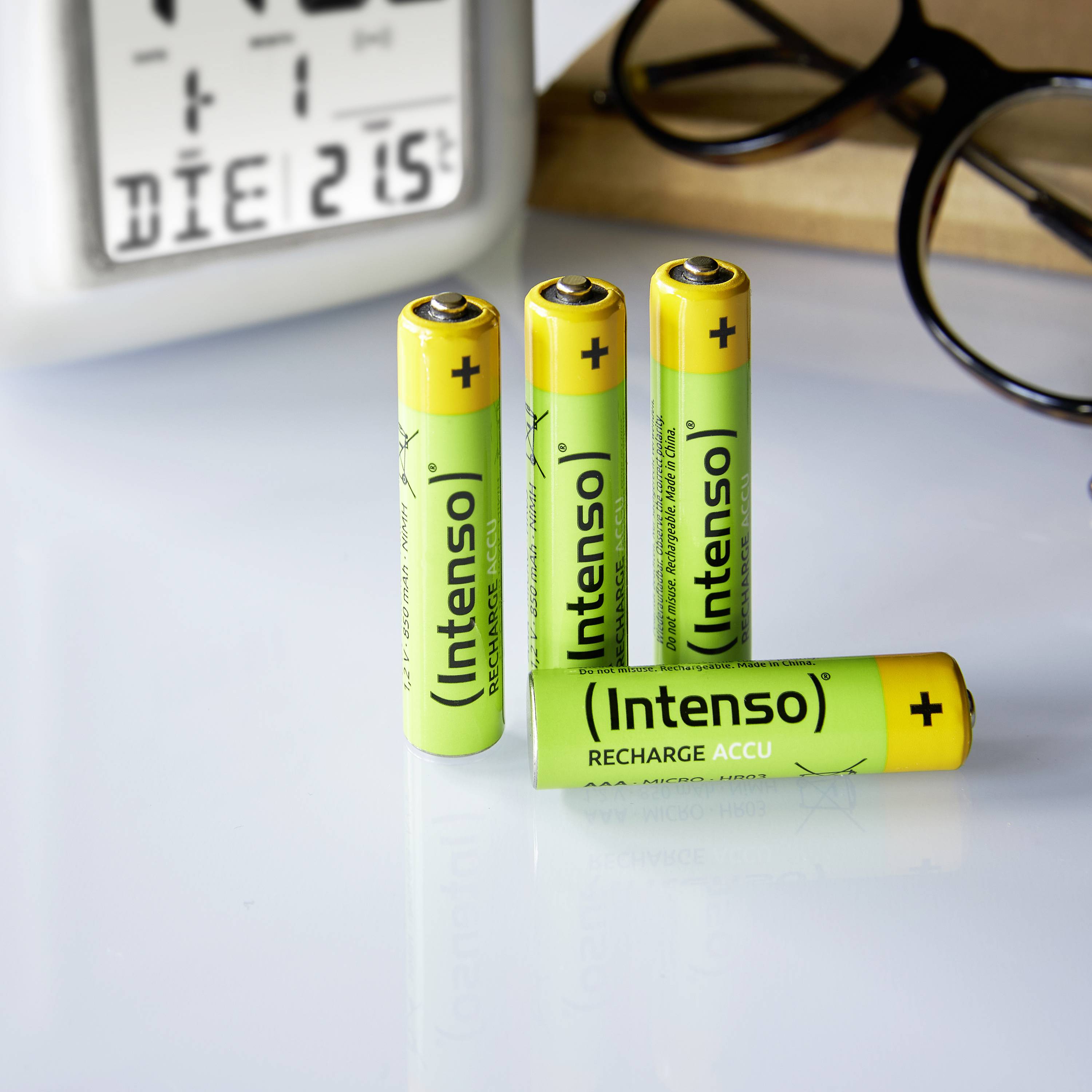 Vier gelbe und grüne AA-Batterien der Marke Intenso stehen auf einem weißen Tisch neben einer digital angezeigten Uhrzeit.