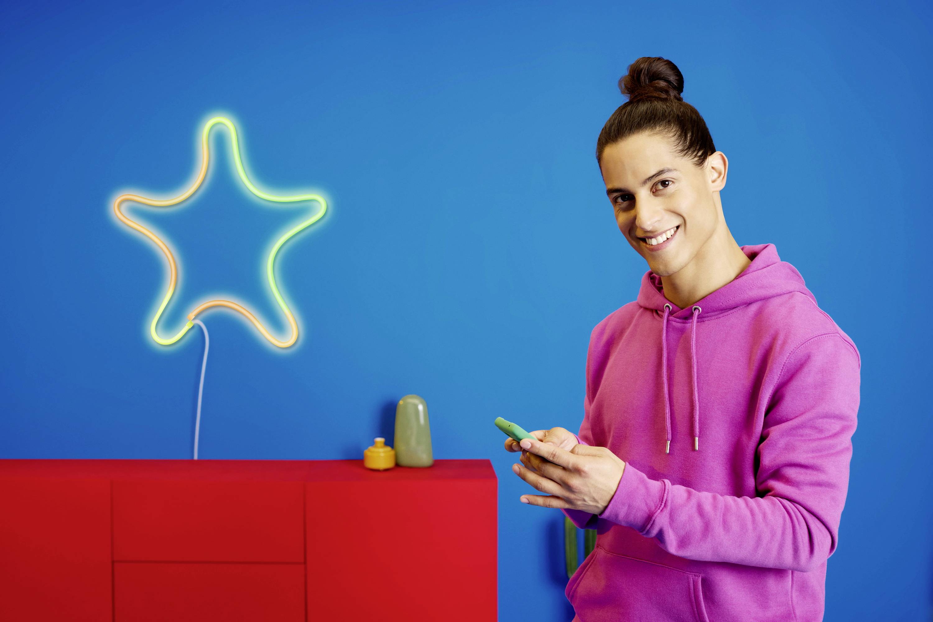Ein lächelnder Mensch in einem pinken Hoodie hält ein Smartphone. Im Hintergrund eine leuchtende Stern-Dekoration vor blauer Wand.