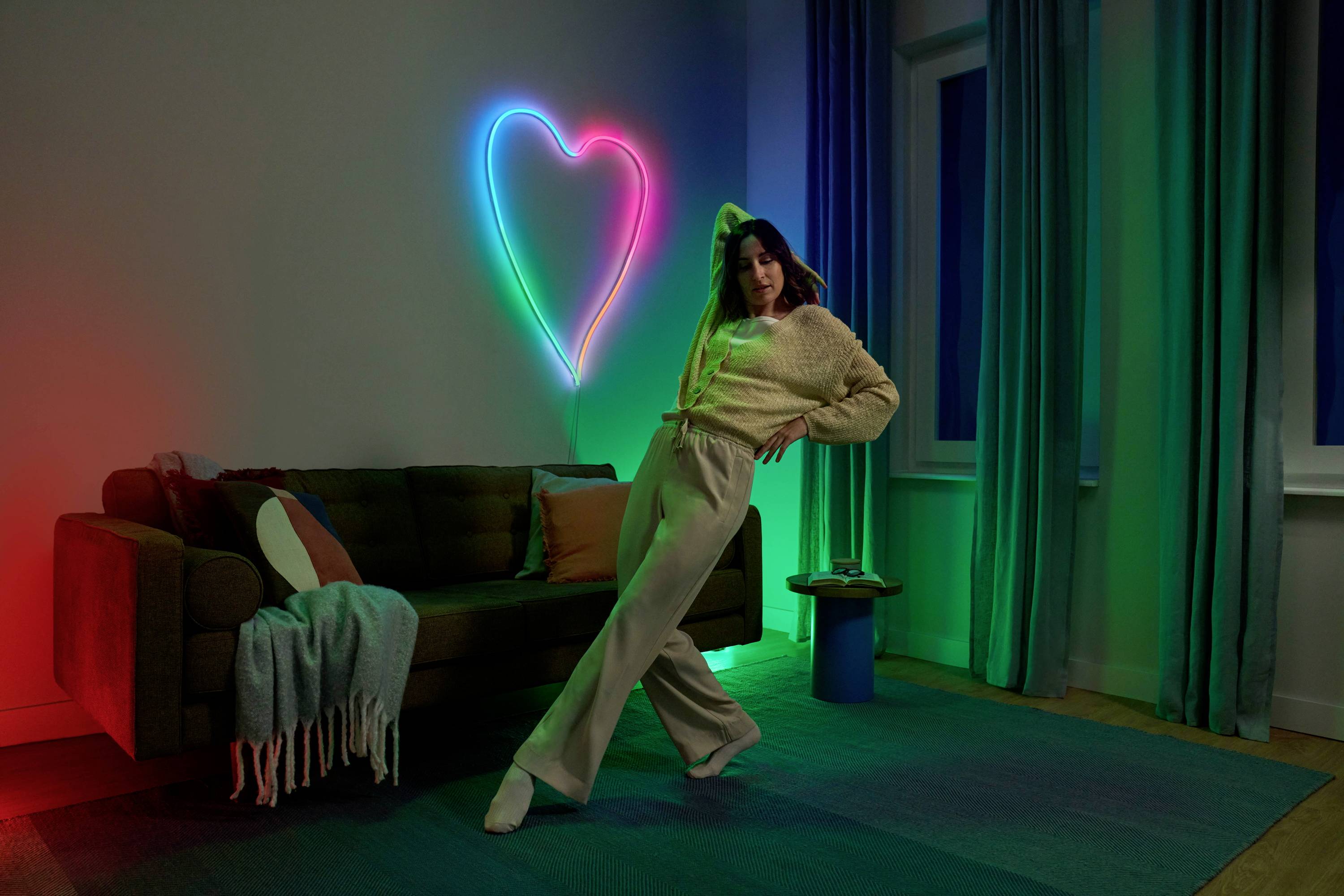 Frau posiert in Wohnzimmer mit Neonherz an der Wand, umgeben von farbigem Licht. Sofa mit Decke und Kissen, Vorhänge an Fenster.