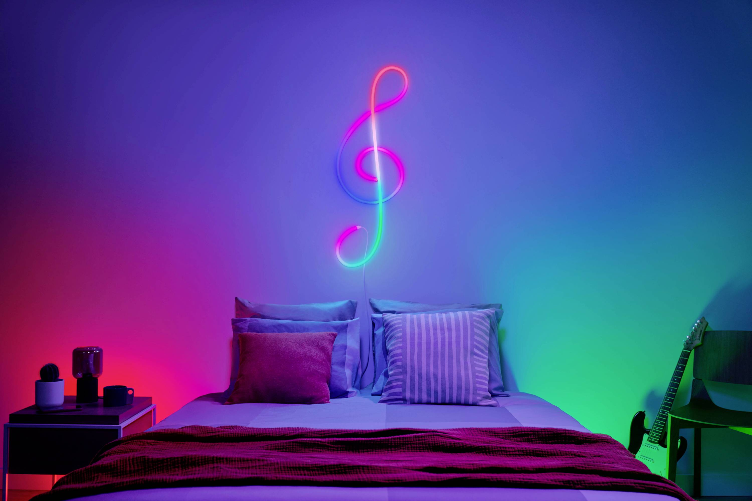 Neonfarbener Notenschlüssel an der Wand über einem Bett mit bunten Kissen, stimmungsvolle Beleuchtung in Blau, Rot und Grün.