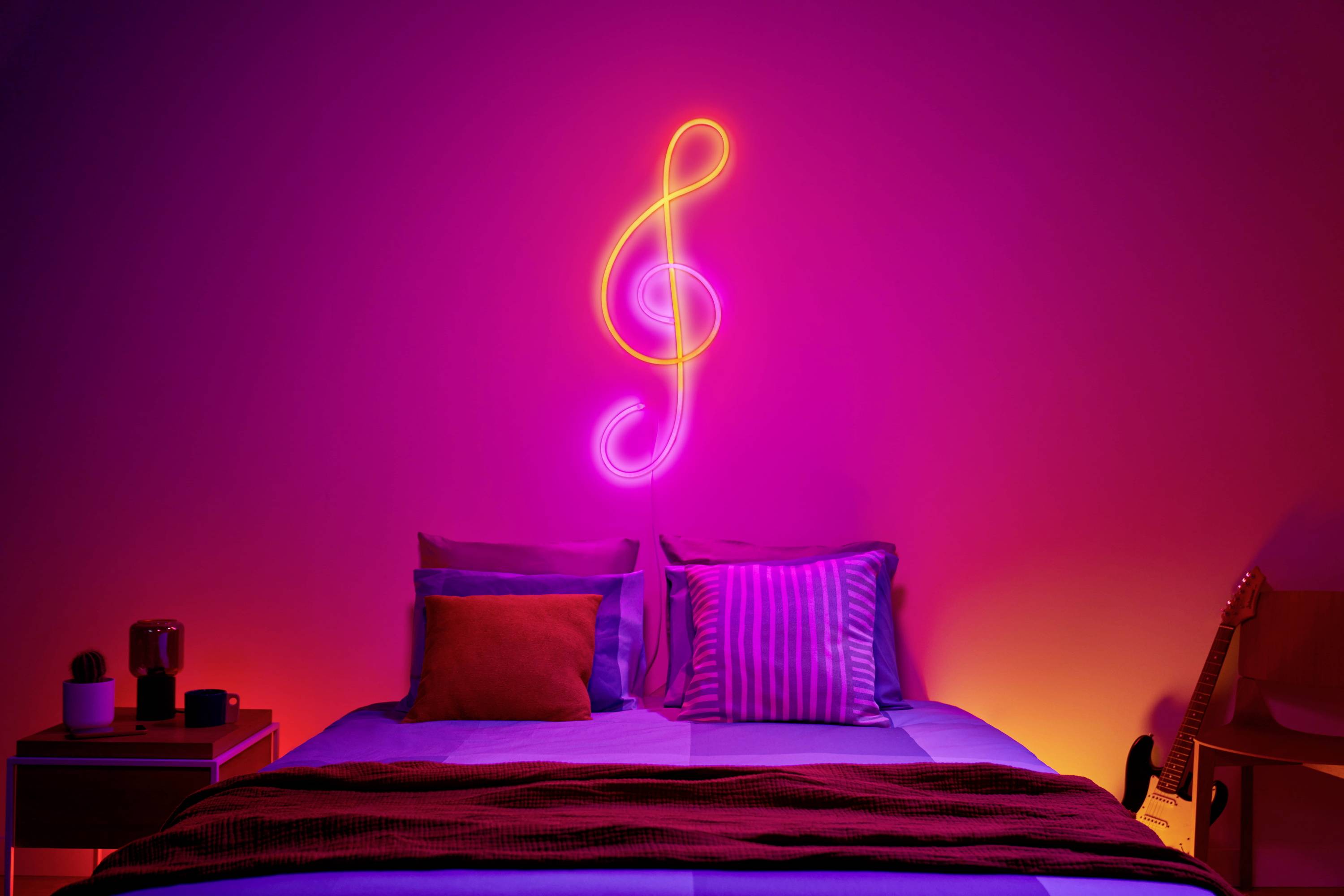 Schlafzimmer mit beleuchtetem Notenschlüssel an der Wand, farbenfrohen Kissen auf dem Bett und Gitarre in der Ecke.