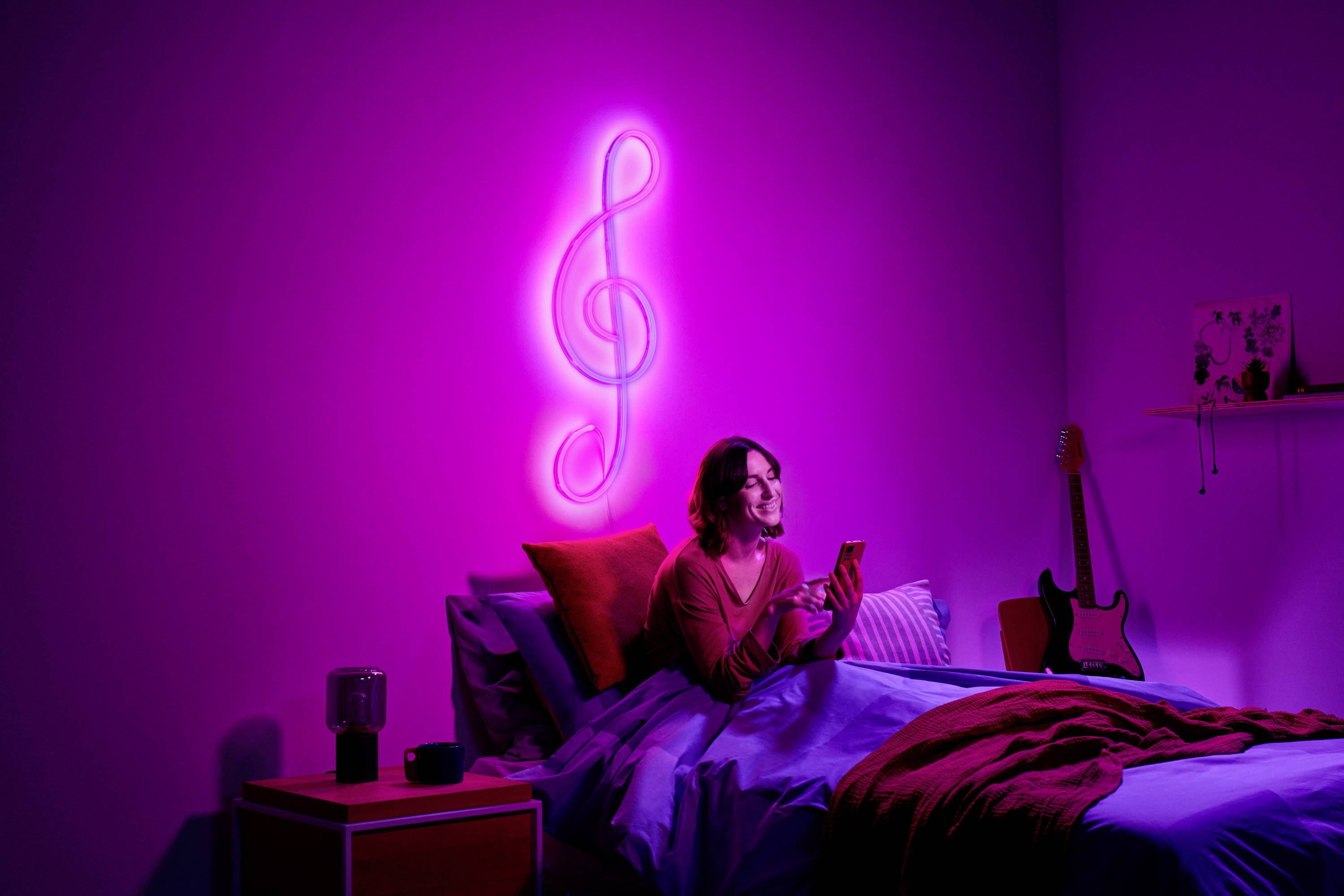 Eine Person sitzt auf einem Bett in einem pink beleuchteten Raum, hält ein Smartphone. An der Wand ein leuchtender Notenschlüssel.
