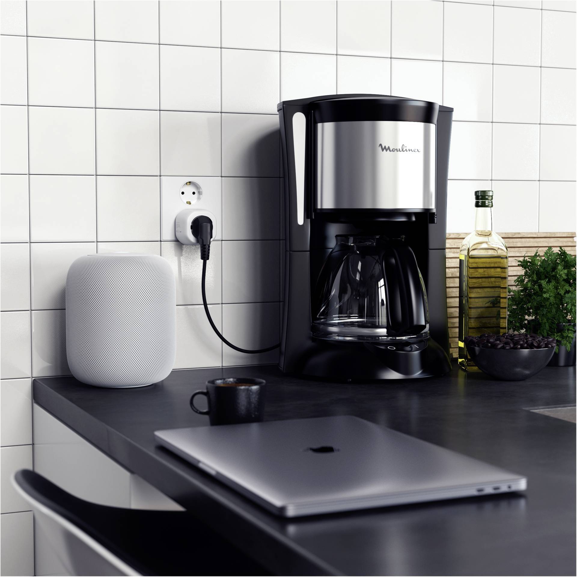 Ein schwarzer Kaffeeautomat steht auf einer Küchenarbeitsplatte neben einem Laptop und einem Lautsprecher, umgeben von Olivenöl und Gewürzen.