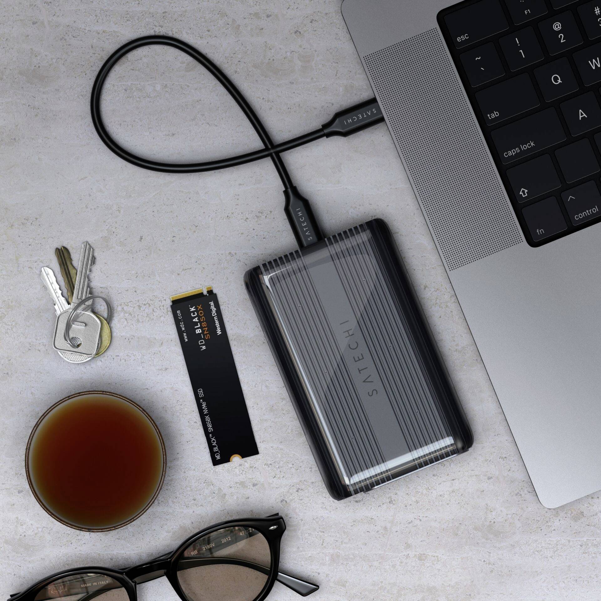 Externe Festplatte angeschlossen an einen Laptop. Daneben liegen Schlüssel, ein Glas Kaffee, eine Brille und ein Speichermodul.