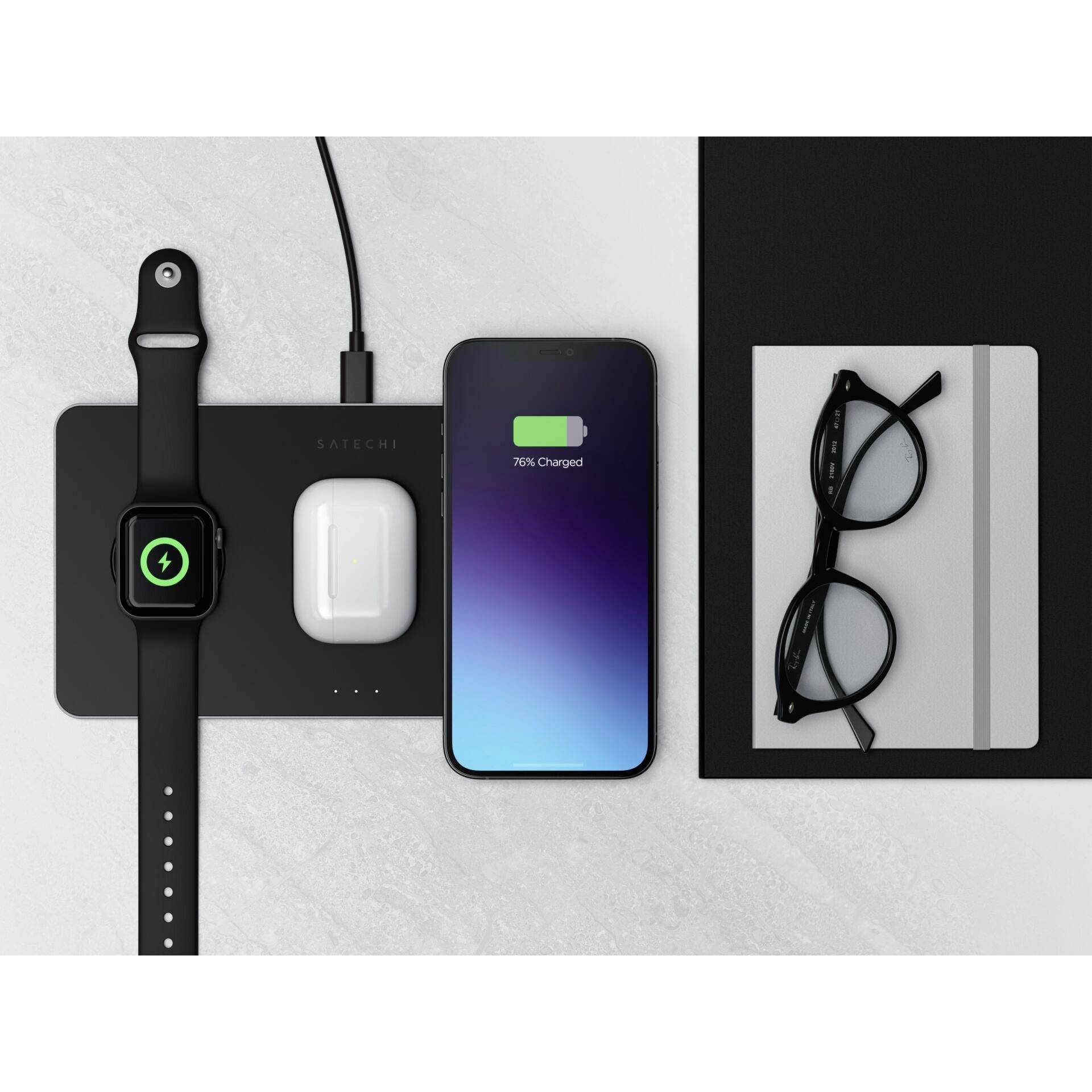 Apple Watch, AirPods und iPhone 12 auf einer kabellosen Ladestation auf einem Tisch; daneben Brillen und ein Notizbuch.