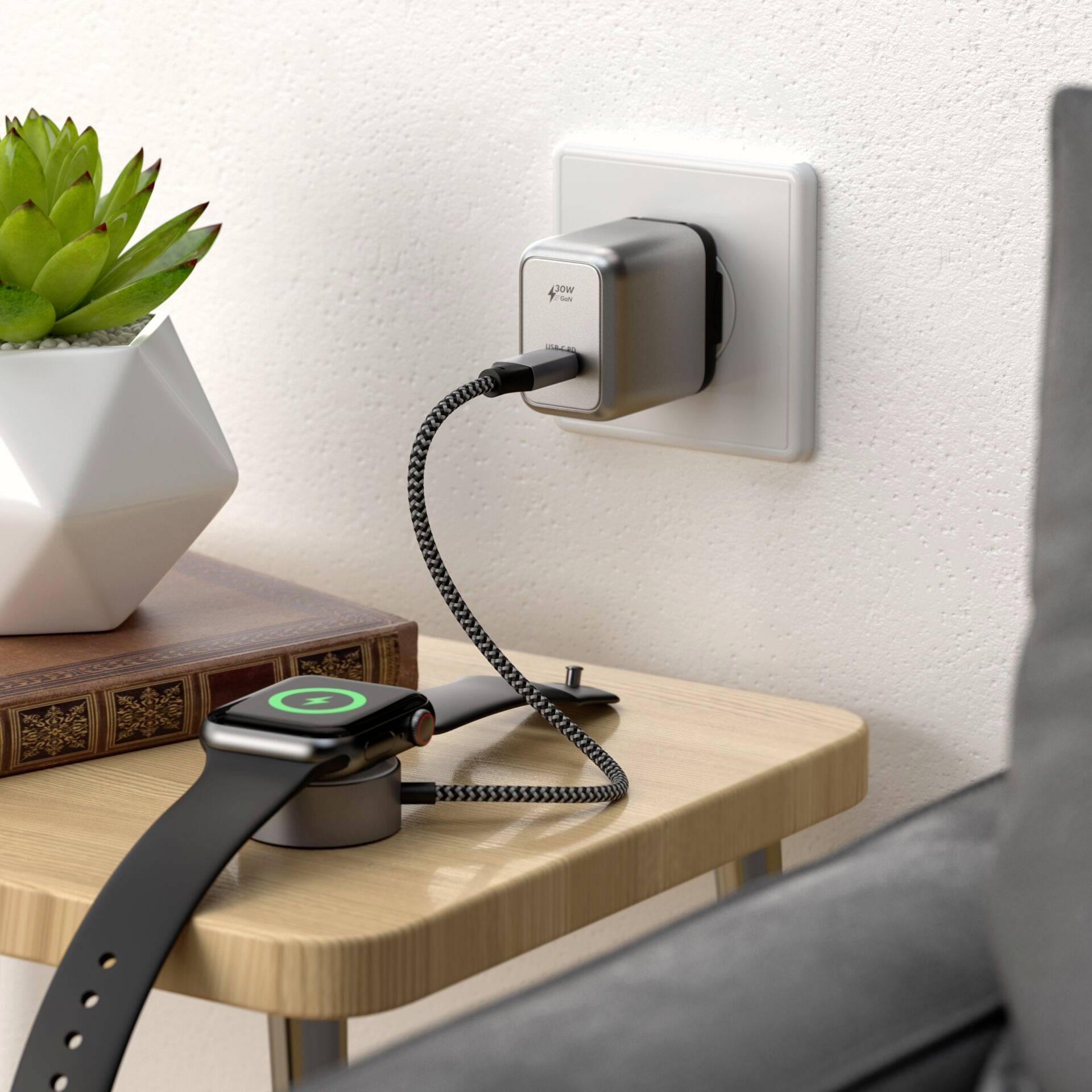 Eine smarte Uhr wird auf einem Tisch aufgeladen. Sie ist mit einem Ladekabel verbunden, das zu einem Adapter an einer Wandsteckdose führt. Neben der Uhr steht eine kleine Pflanze.