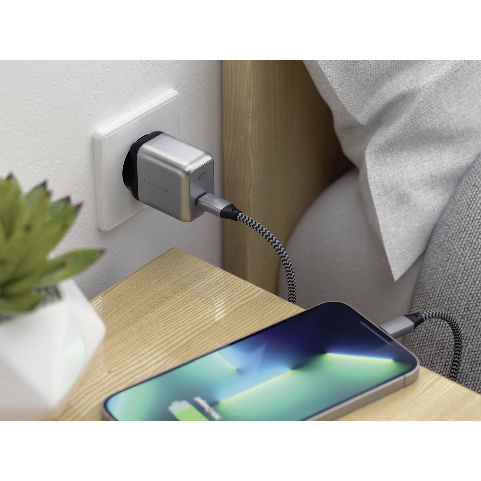 Ein Smartphone wird mittels eines geflochtenen Kabels an eine Steckdose aufgeladen. Das Telefon liegt auf einem Holztisch neben einer Pflanze.