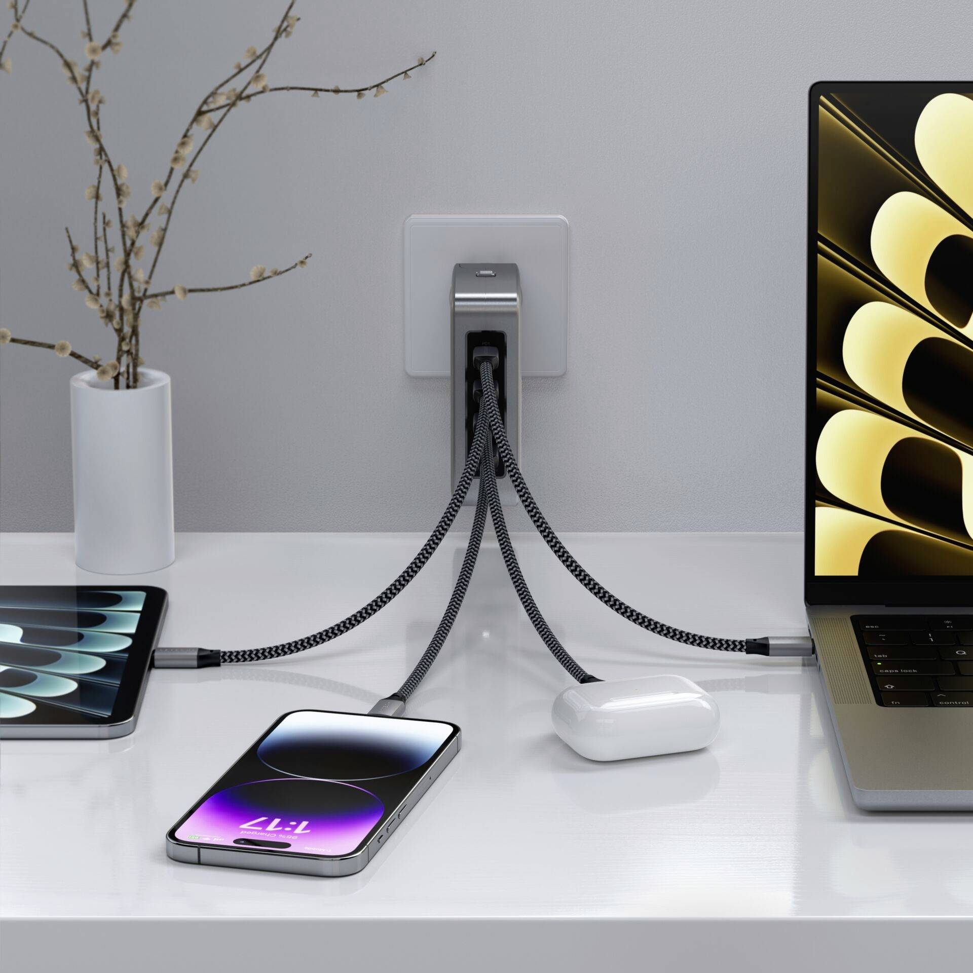 Smartphone und Laptop sind an einer Mehrfachsteckdose angeschlossen, die an eine Wandsteckdose angeschlossen ist. Links steht eine Vase mit Zweigen.