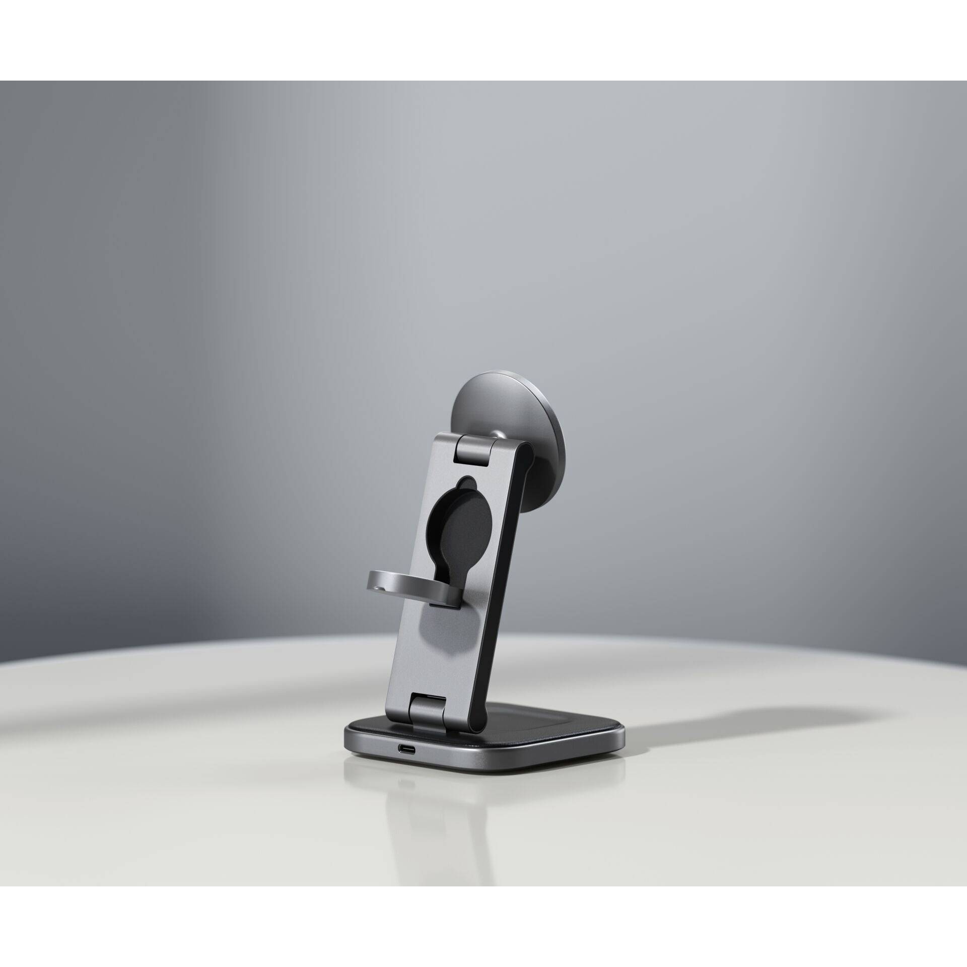 Ein grauer Smartphone-Ständer auf einem weißen Tisch, in einem minimalistischen Raum platziert. Der Ständer ist klappbar.