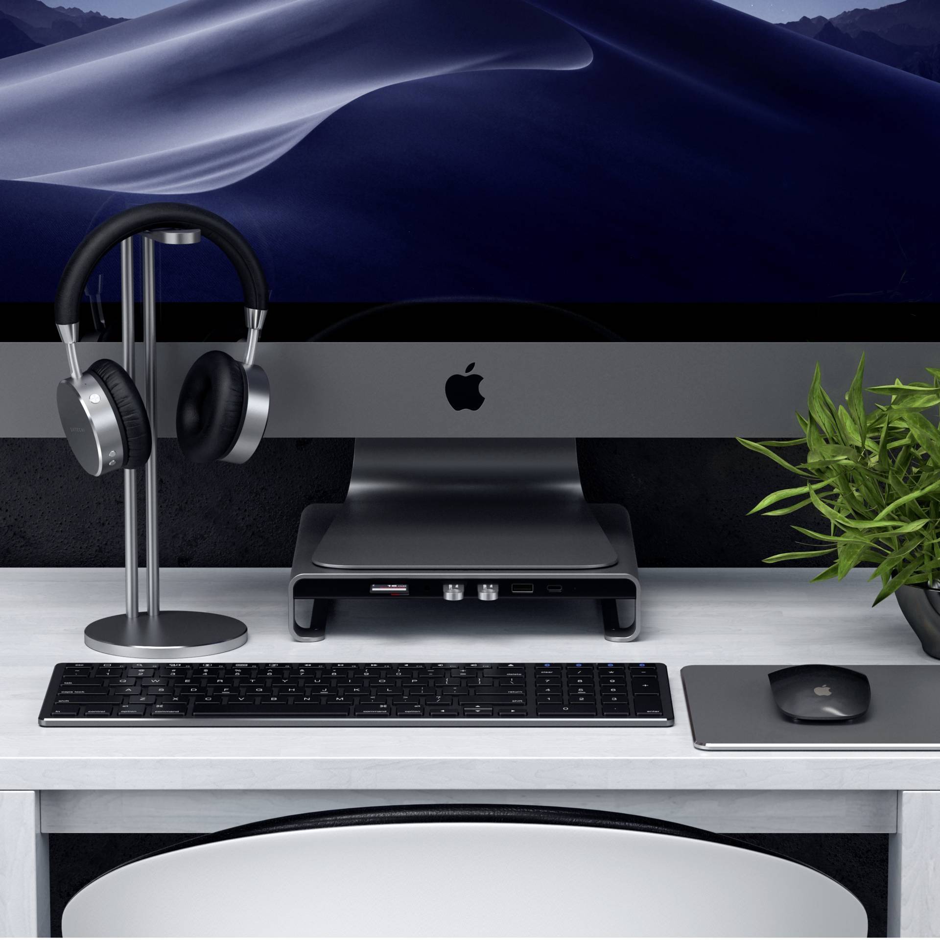 Ein Schreibtisch mit einem iMac, einem Paar Kopfhörern auf einem Ständer, einer Tastatur und einer Maus. Eine grüne Pflanze steht rechts.