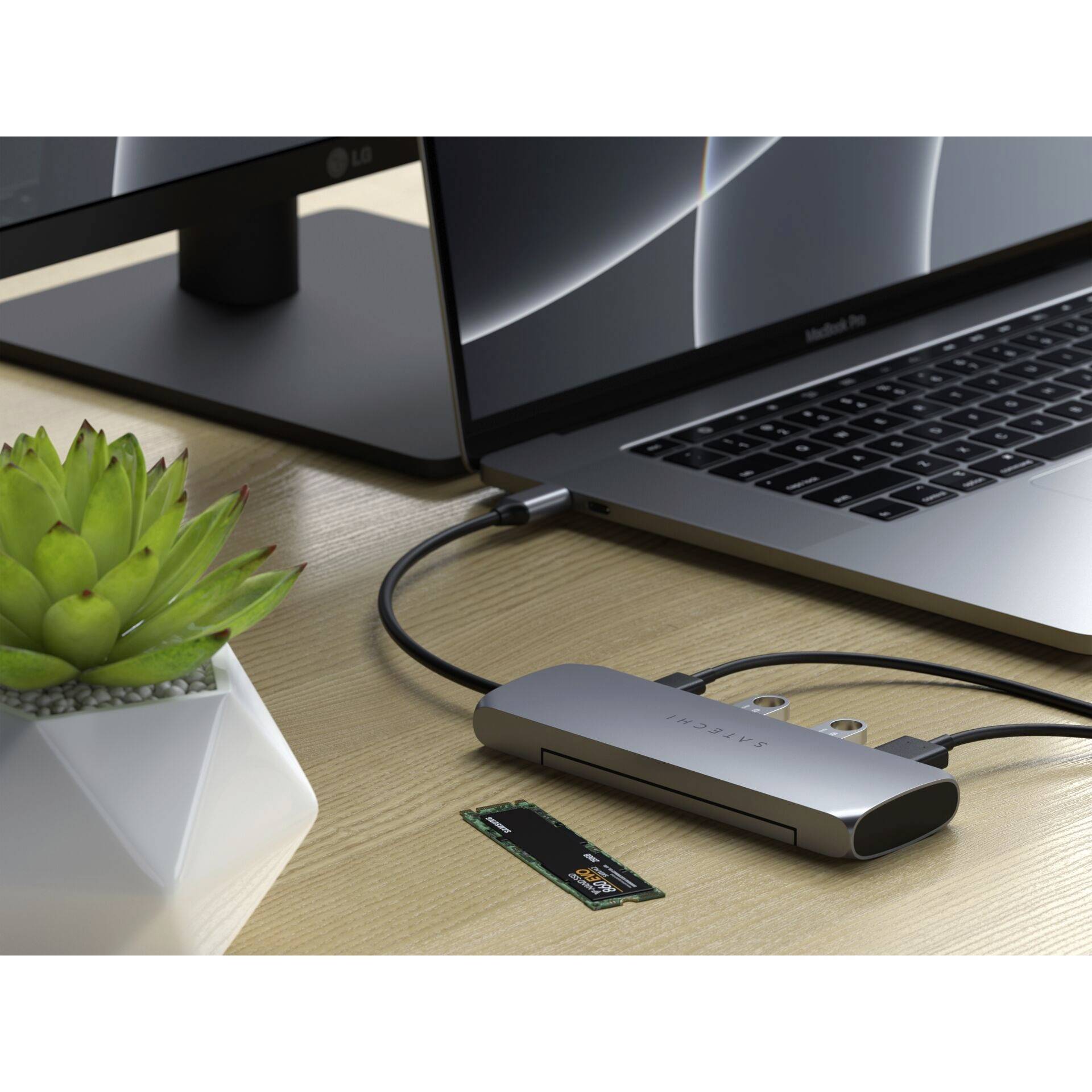 Ein Laptop ist mit einem USB-C-Hub verbunden, der an einen Monitor angeschlossen ist. Eine Pflanze steht daneben auf dem Tisch.