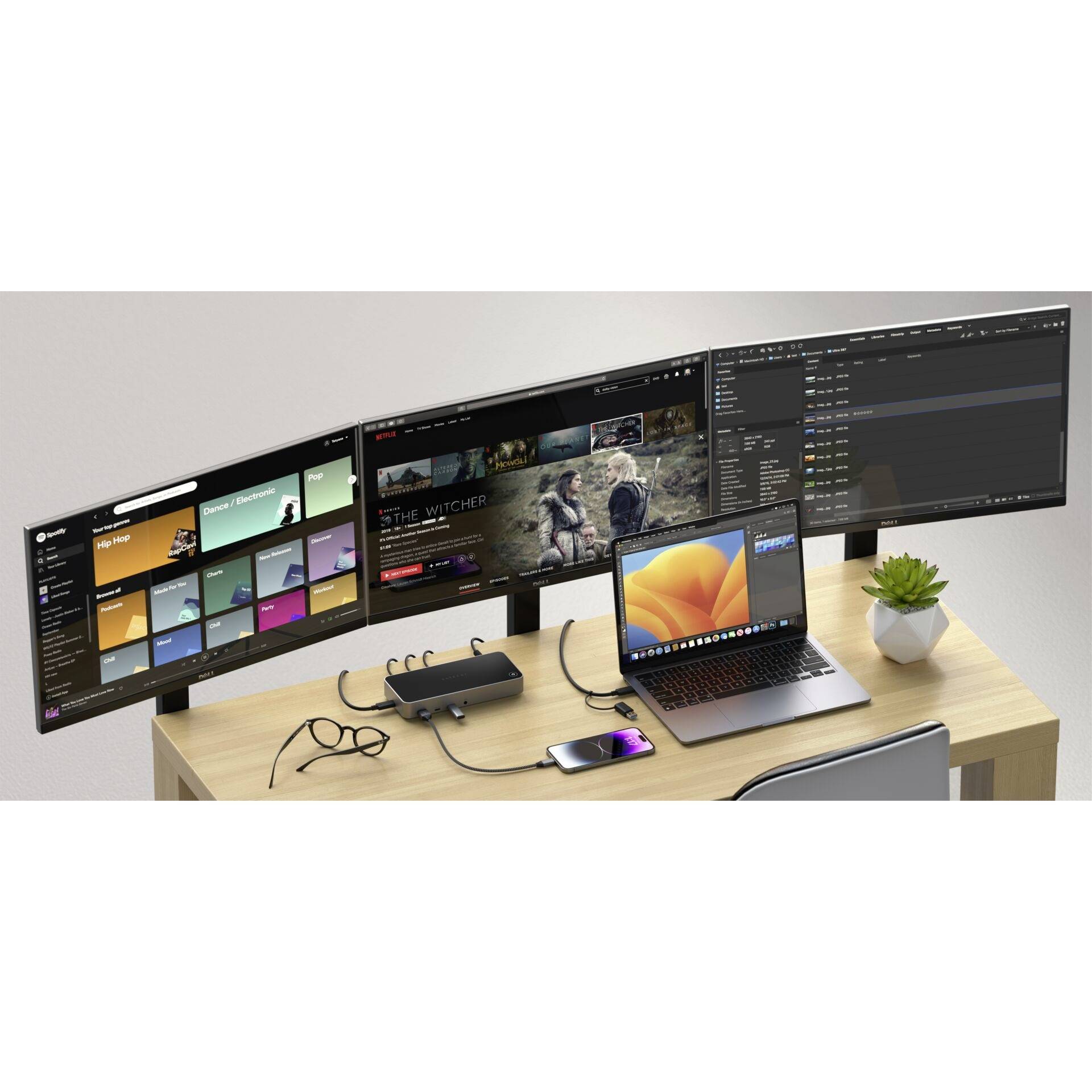 Ein Schreibtisch mit drei Monitoren zeigt verschiedene Anwendungen, ein Laptop und ein Smartphone sind angeschlossen, daneben eine Pflanze.