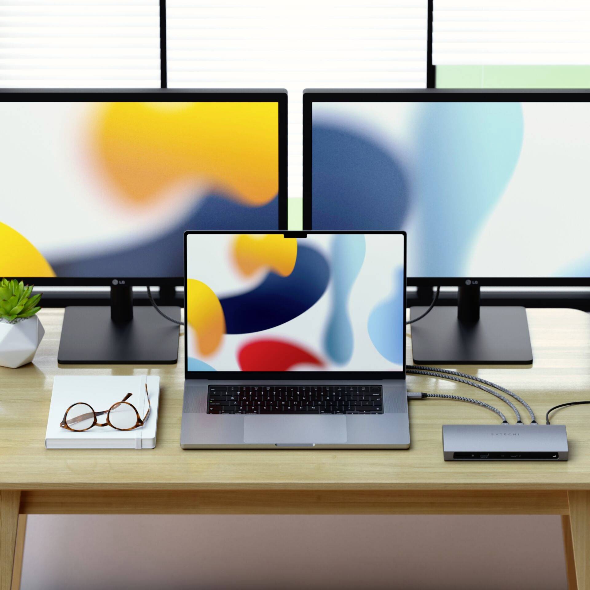 Ein Schreibtisch mit zwei Monitoren und einem Laptop, verbunden über eine Dockingstation; Bücher, Brille und Topfpflanze daneben.