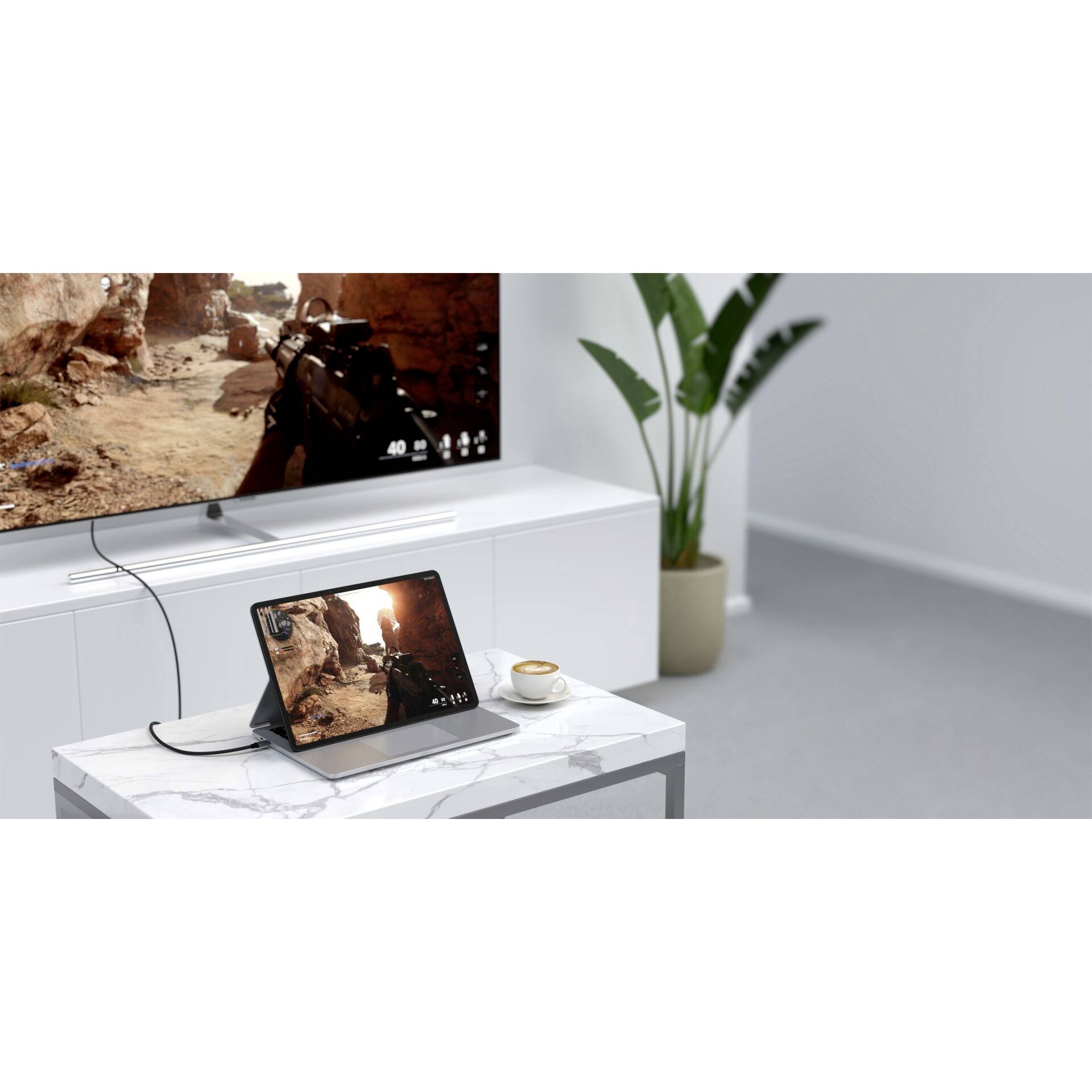 Tablet und Fernseher auf weißem Tisch; das Tablet zeigt ein Videospiel, das auch auf dem großen Bildschirm wiedergegeben wird. Im Hintergrund steht eine Pflanze.