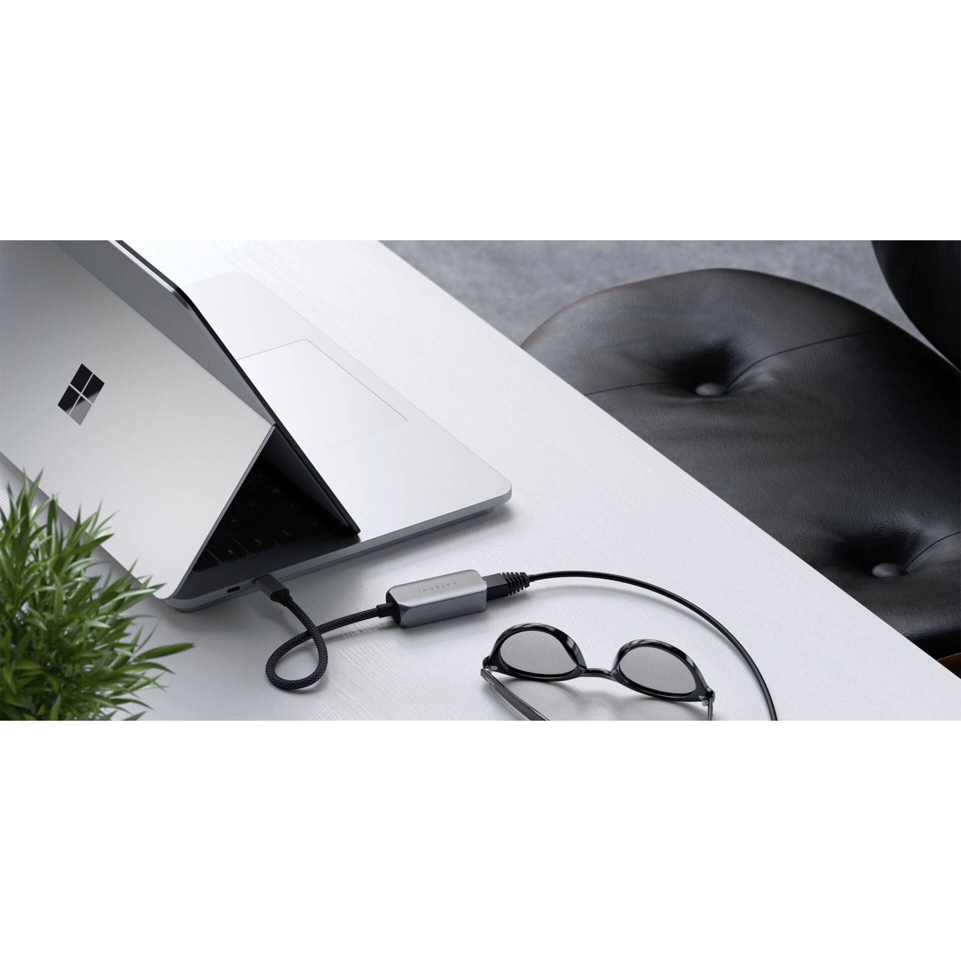 Ein Laptop ist an einen Adapter auf einem Schreibtisch angeschlossen. Daneben liegen eine Brille und eine Topfpflanze.