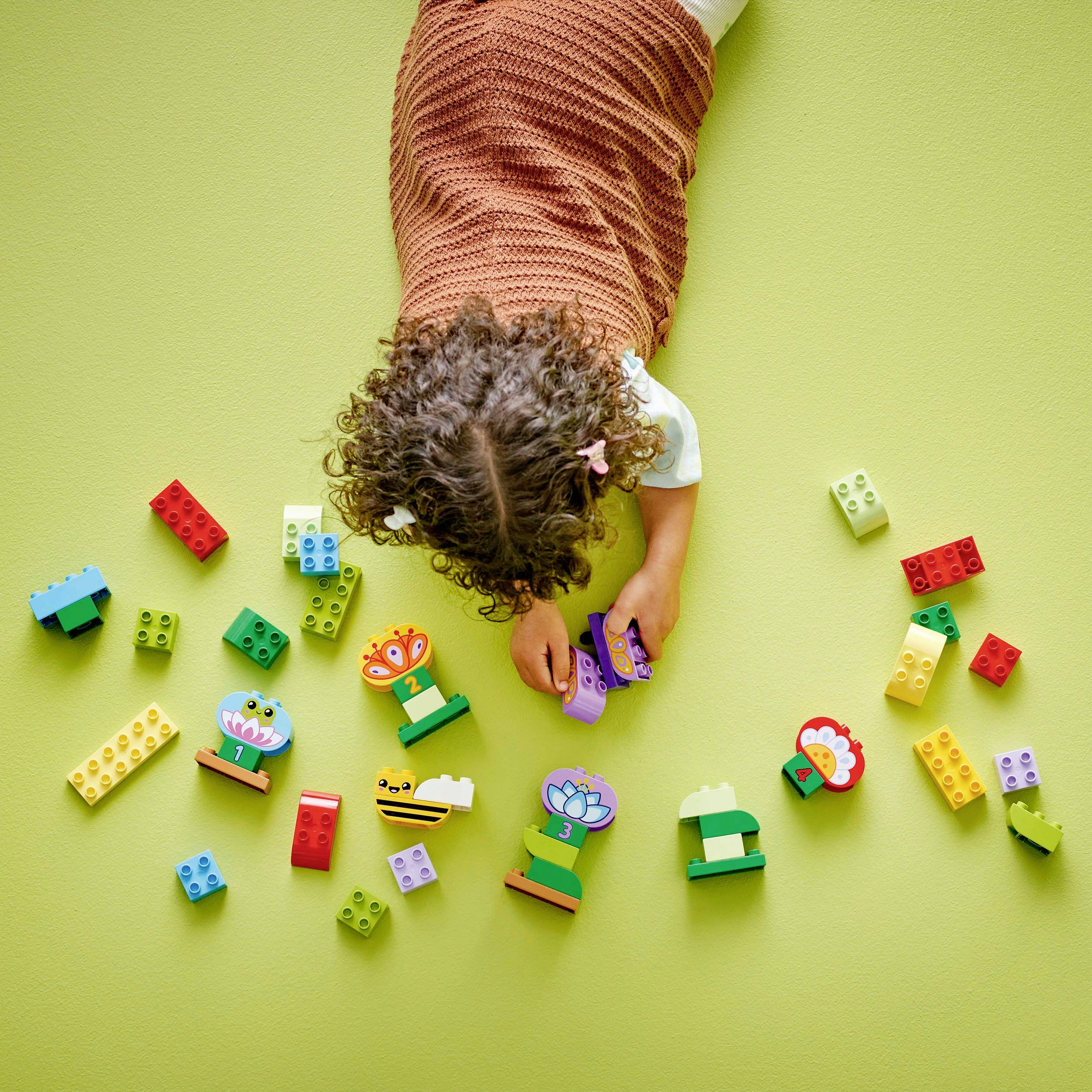 Ein Kind spielt auf einem grünen Teppich mit bunten Bauklötzen und Blumenfiguren. Das Kind trägt ein braunes Oberteil.