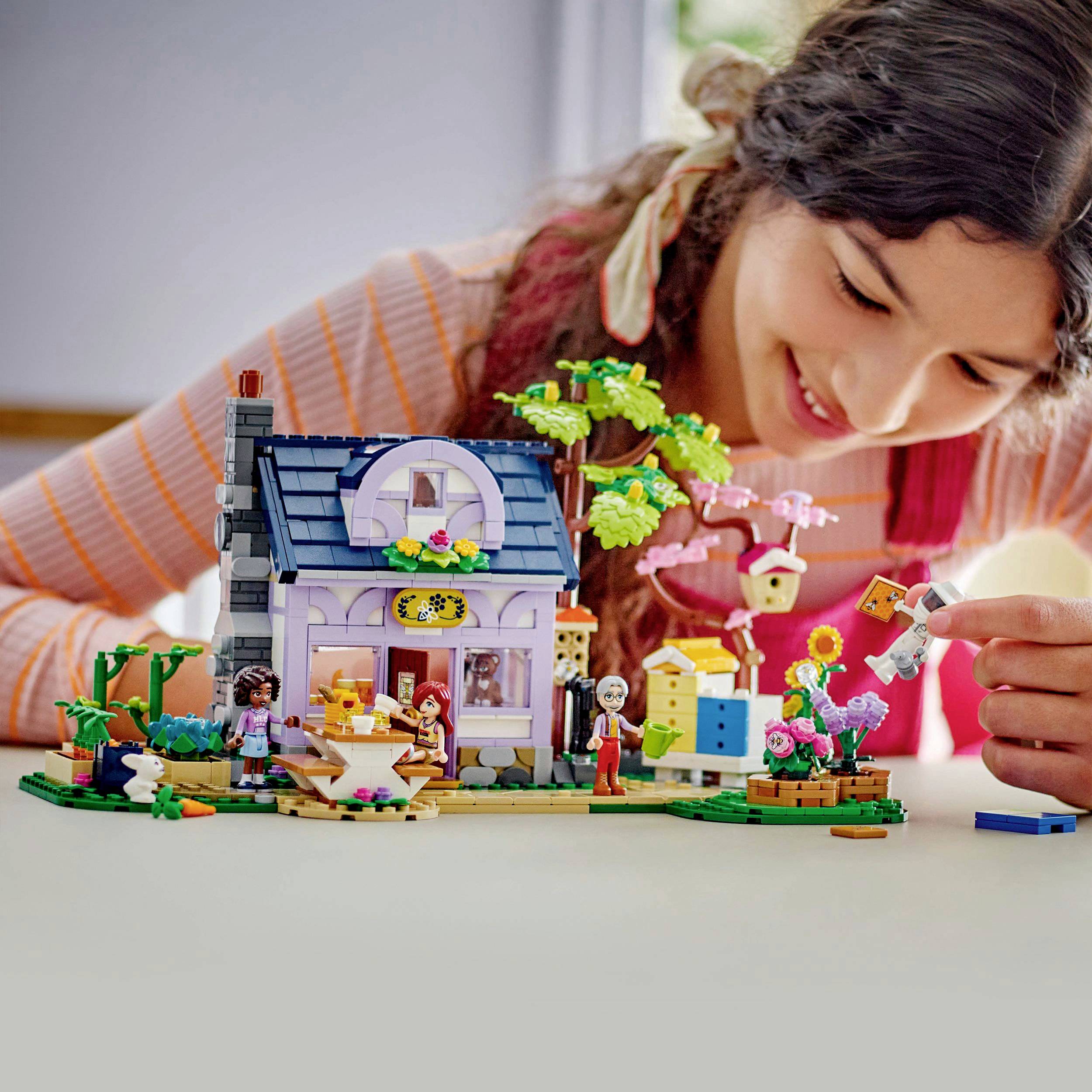 Ein Mädchen spielt mit einem LEGO-Set, das ein Haus mit Garten darstellt. Das Set enthält Figuren, Tiere und Pflanzen und vermittelt eine fröhliche, kreative Atmosphäre.