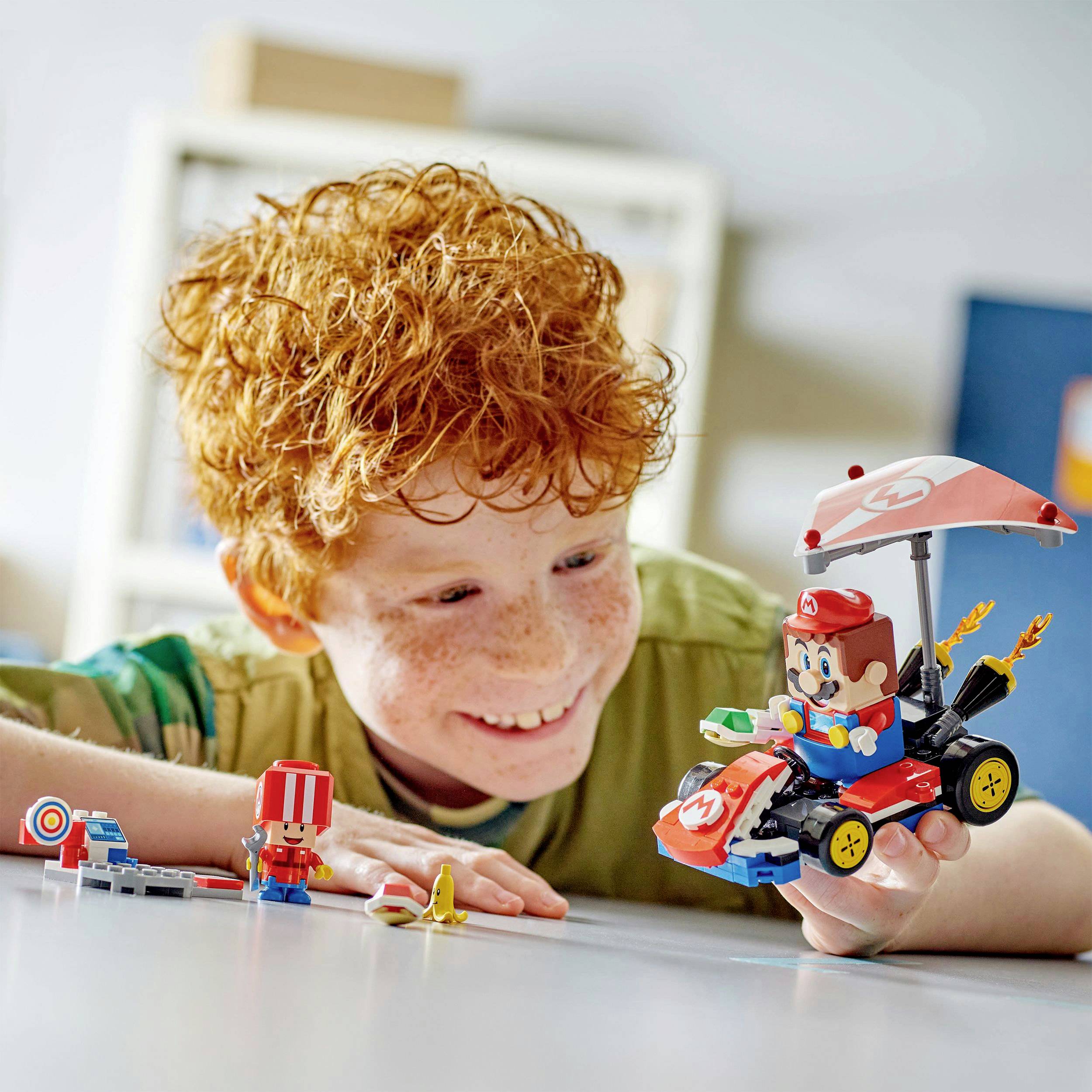 Ein Kind mit roten Haaren spielt begeistert mit einem selbstgebauten Lego-Rennwagen, der von einer bekannten Videospielfigur inspiriert ist.