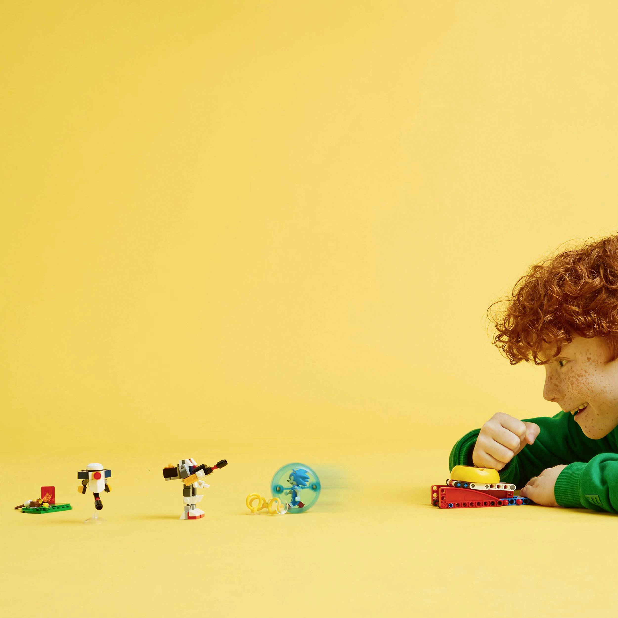 Ein Kind mit lockigen Haaren spielt freudig mit baukästenartigen Spielzeugen auf einem gelben Tisch vor einem gelben Hintergrund.