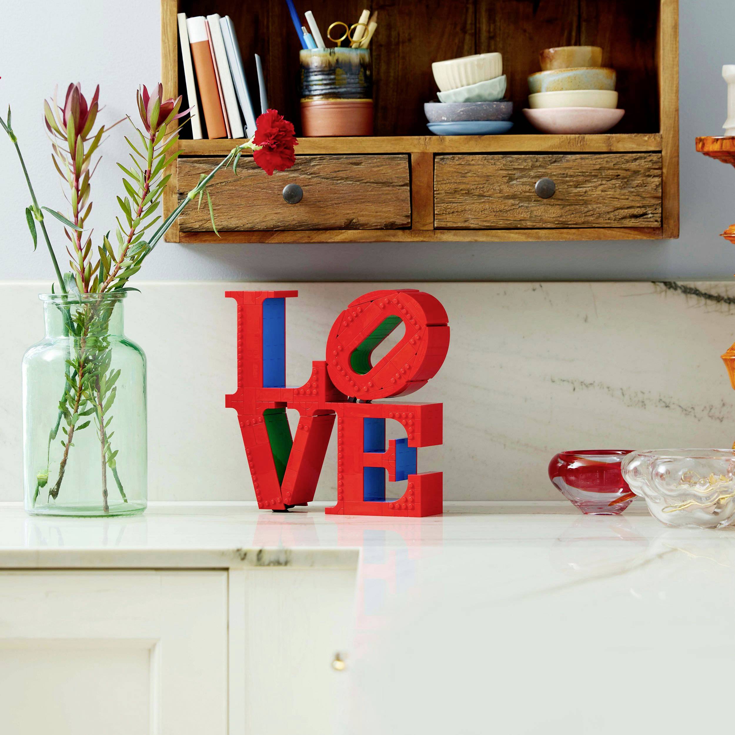 Ein Küchenregal mit Geschirr. Auf der Arbeitsplatte steht eine Glasvase mit Blumen und die Buchstaben 'LOVE' in Rot neben einer roten Schale.