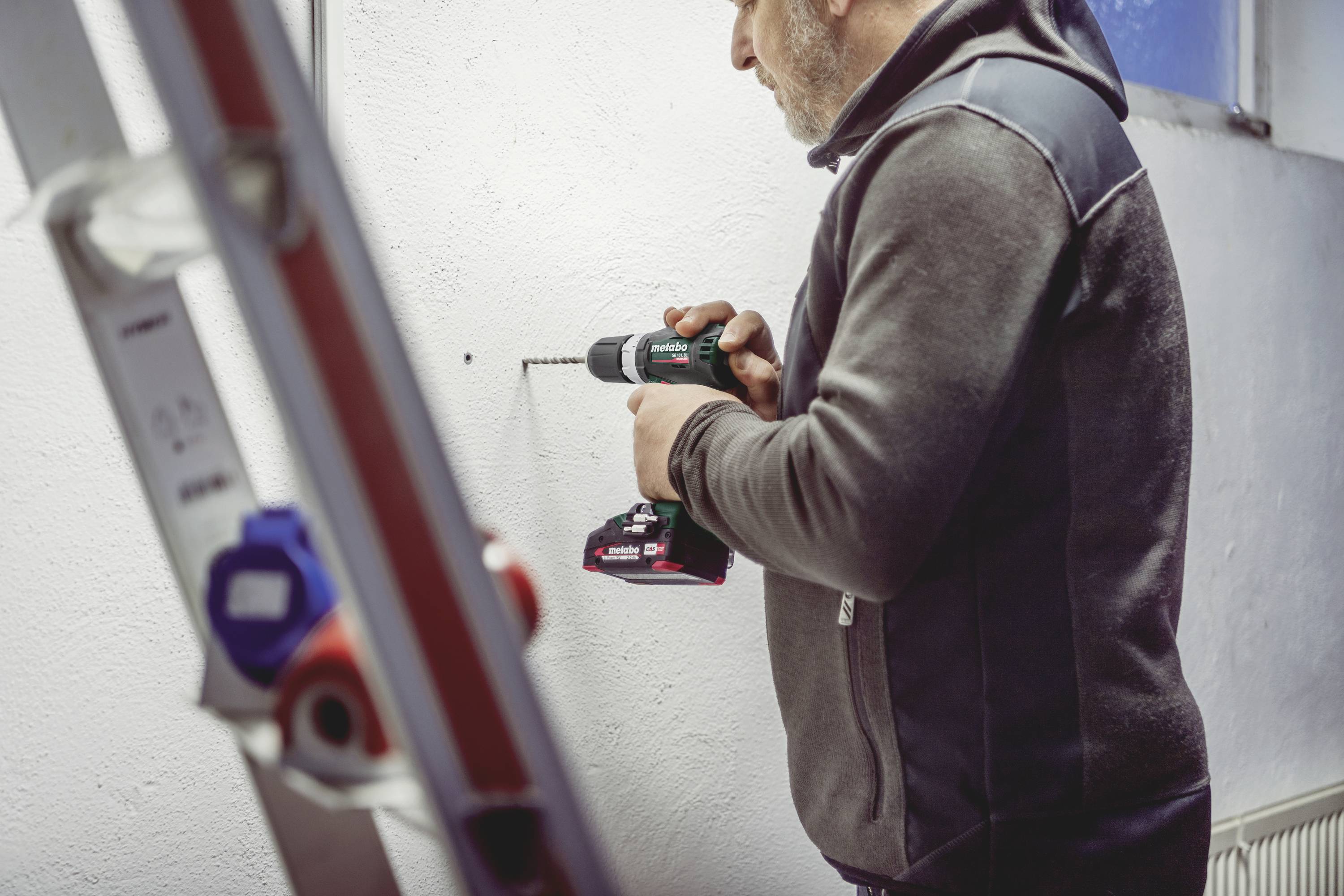 Mann bohrt mit einer elektrischen Bohrmaschine ein Loch in die weiße Wand. Eine Leiter steht im Vordergrund.