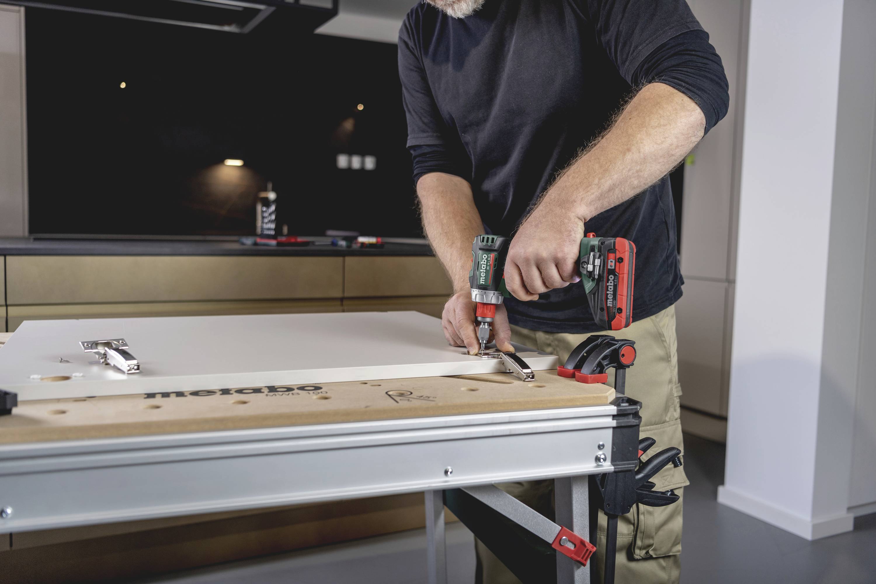 Eine Person befestigt mit einer elektrischen Bohrmaschine ein Scharnier an einer weißen Tür in einer modernen Küche.