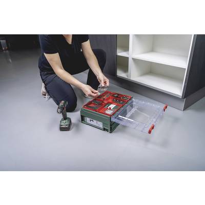 Eine Person kniet auf dem Boden und sortiert Schrauben in einer grünen Werkzeugbox neben einem Akkuschrauber und einem Regal.