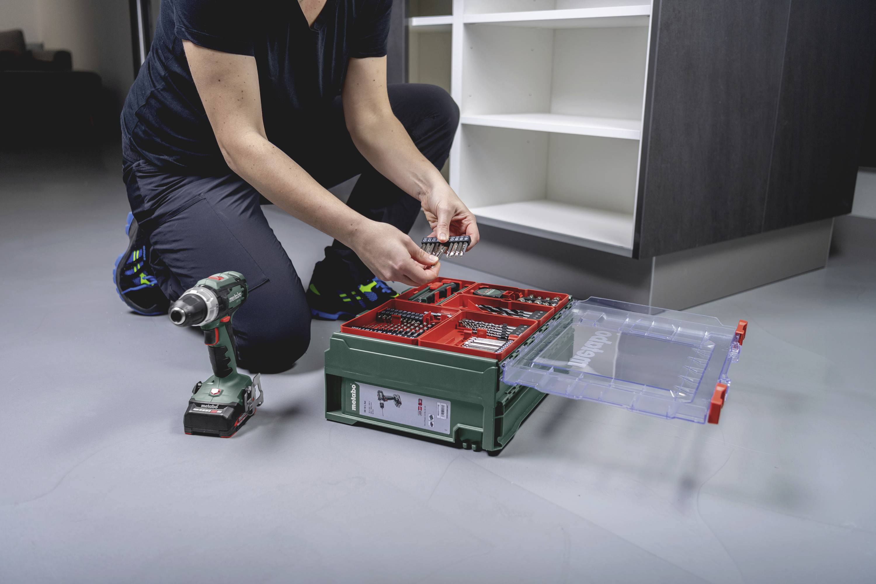 Eine Person kniet auf dem Boden und sortiert Schrauben in eine Werkzeugbox neben einer Bohrmaschine vor einem Schrank.